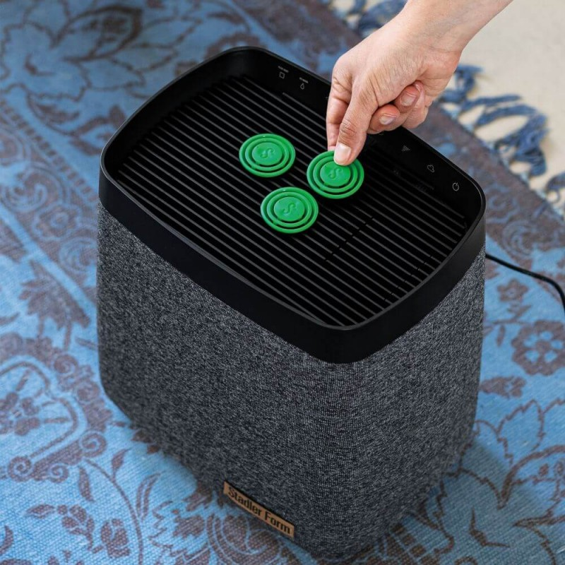 Osoba umieszczająca zielone dyski filtrujące na szarym oczyszczaczu powietrza. Niebieski dywan w tle.