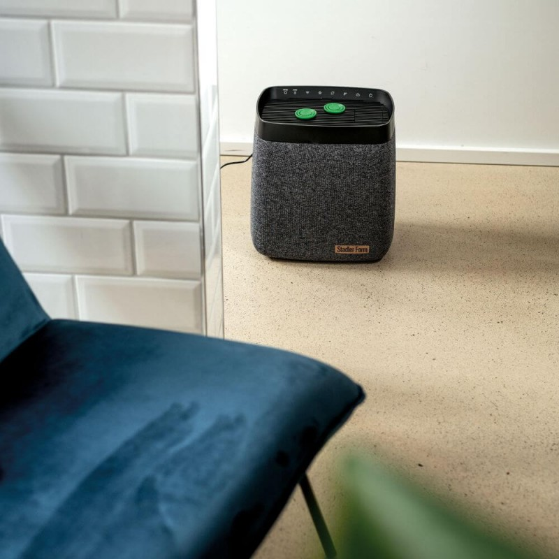 Oczyszczacz powietrza z szarą tkaniną, zielonymi przyciskami i niebieskim krzesłem na pierwszym planie.