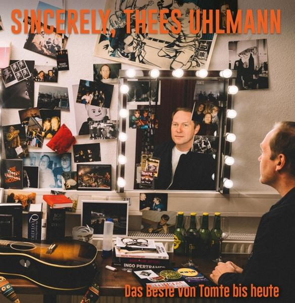 Mann im Spiegel, umgeben von Fotos und Lichtern. Backstage-Szene mit Gitarre und Getränken.