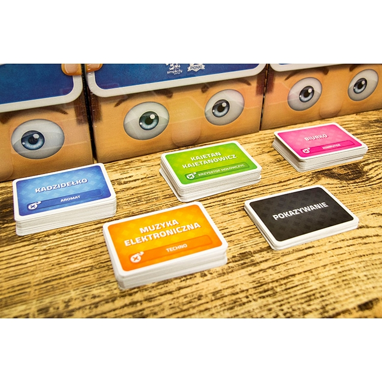 Karty z gry planszowej, pokazujące twarze i różne słowa po polsku, na drewnianej powierzchni.