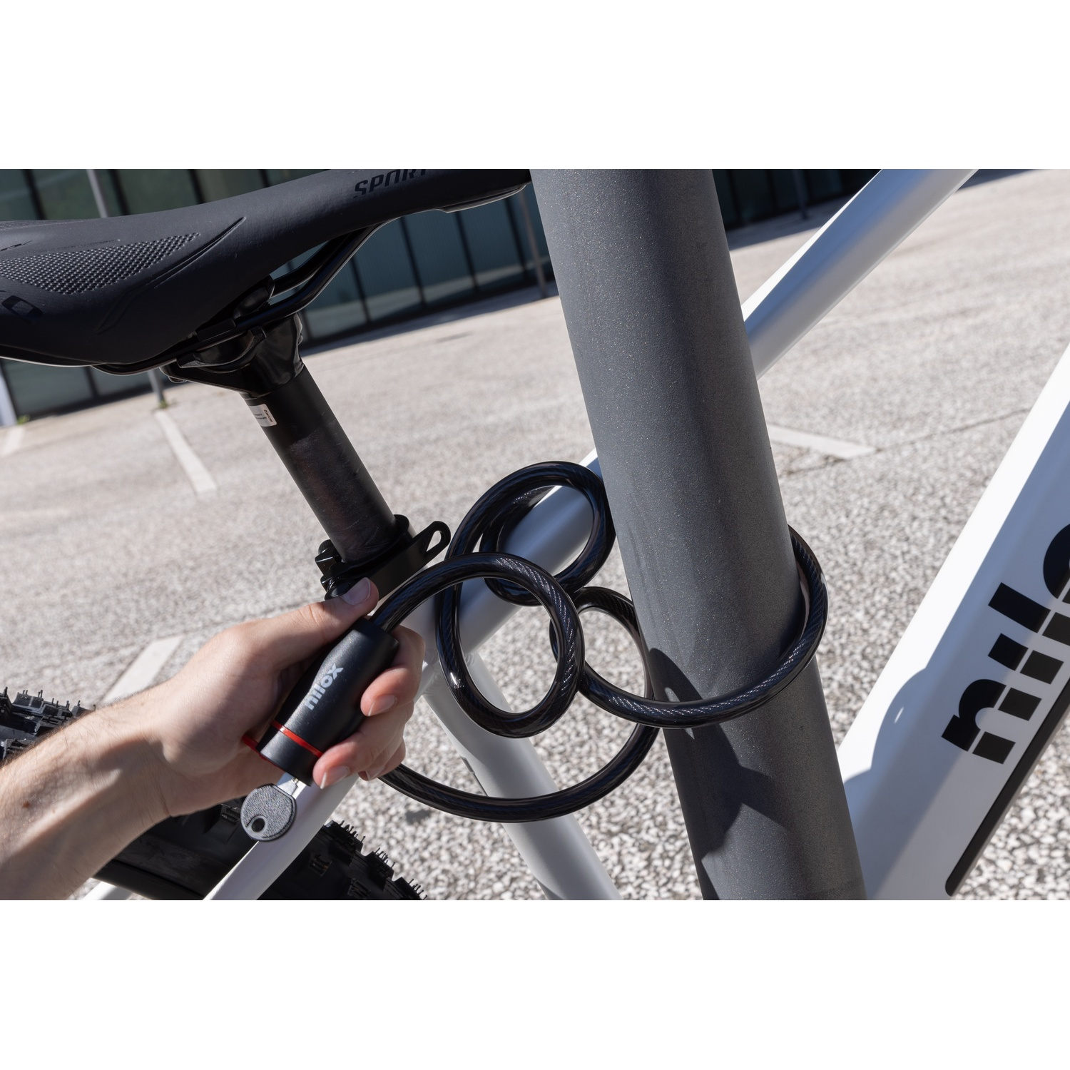 Una mano blocca un lucchetto nero per bici attorno a un palo grigio di una bicicletta. La bici è bianca.
