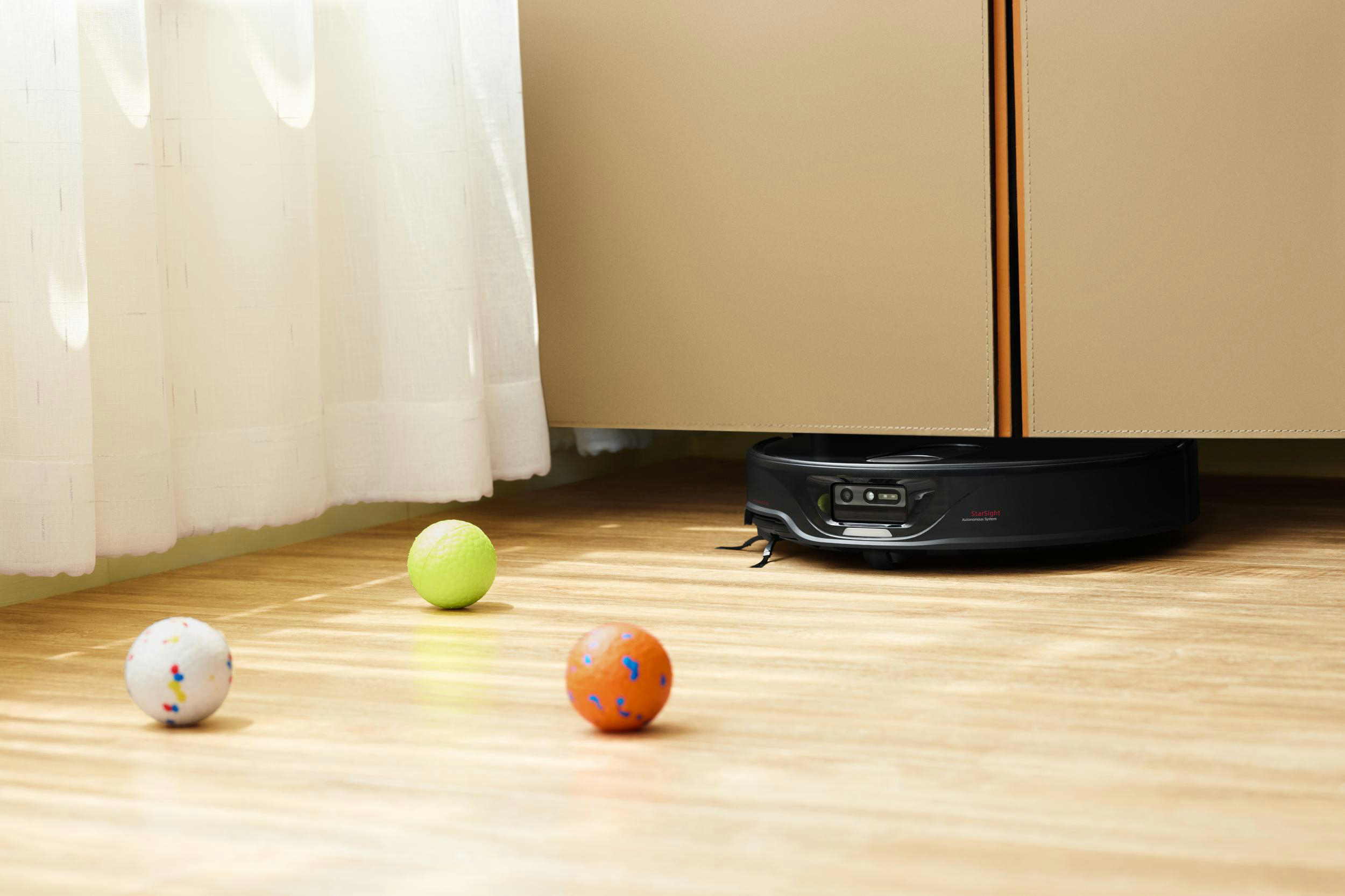Ein Roboter-Staubsauger reinigt einen Holzboden mit drei bunten Bällen.