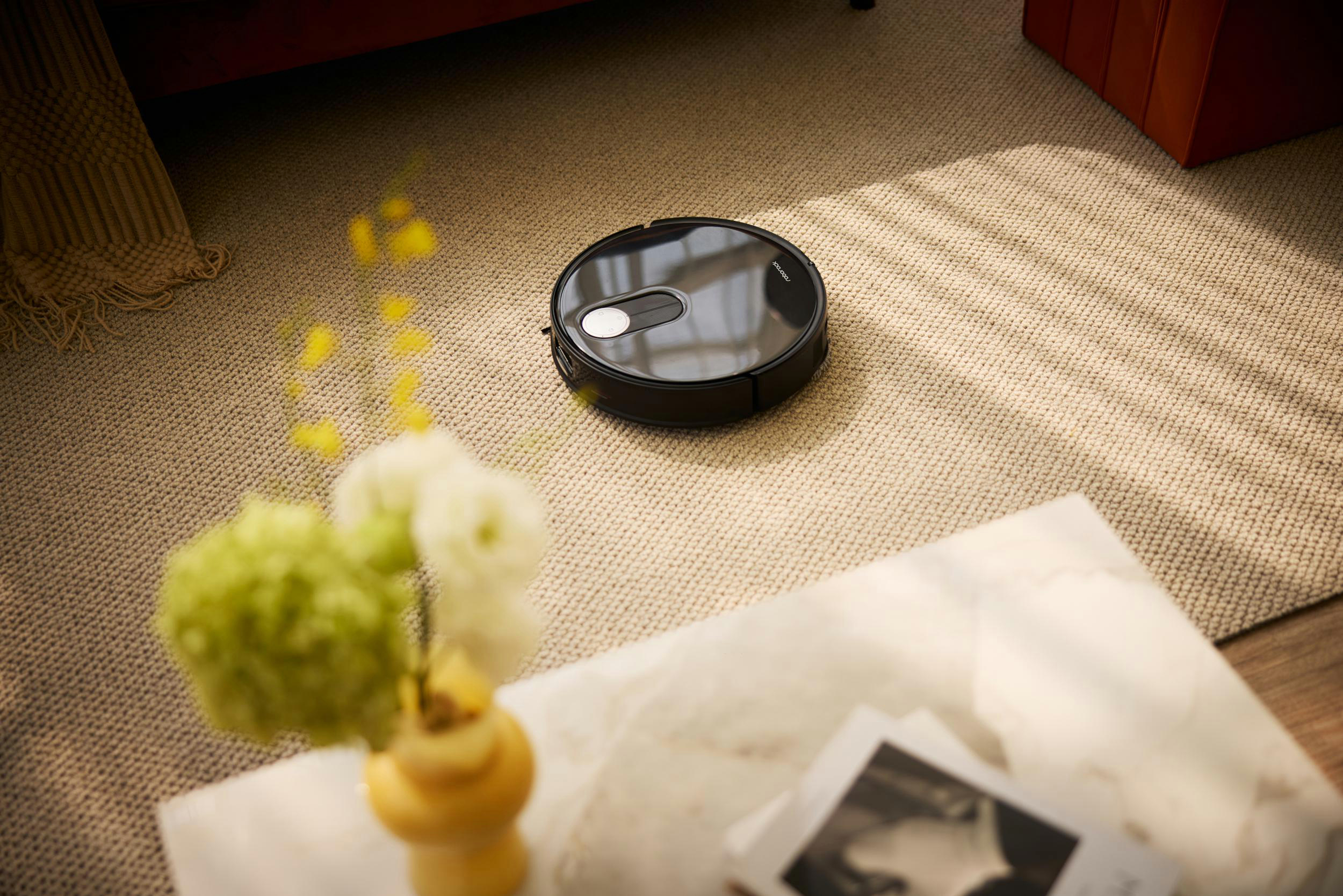 Ein schwarzer Roboter-Staubsauger auf einem Teppich neben einem Tisch mit Blumen und Zeitschriften.