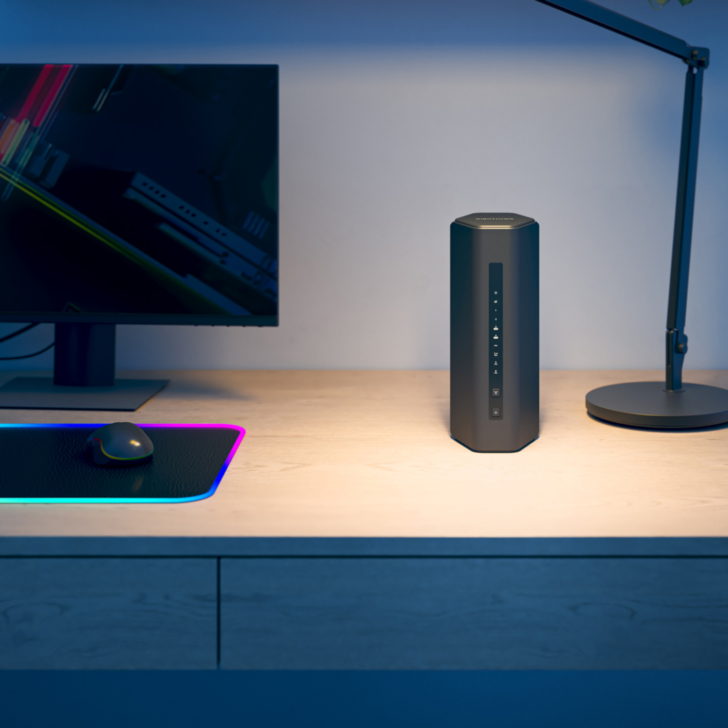 Een zwarte router op een houten bureau. Een computermonitor en een bureaulamp staan ook op het bureau.