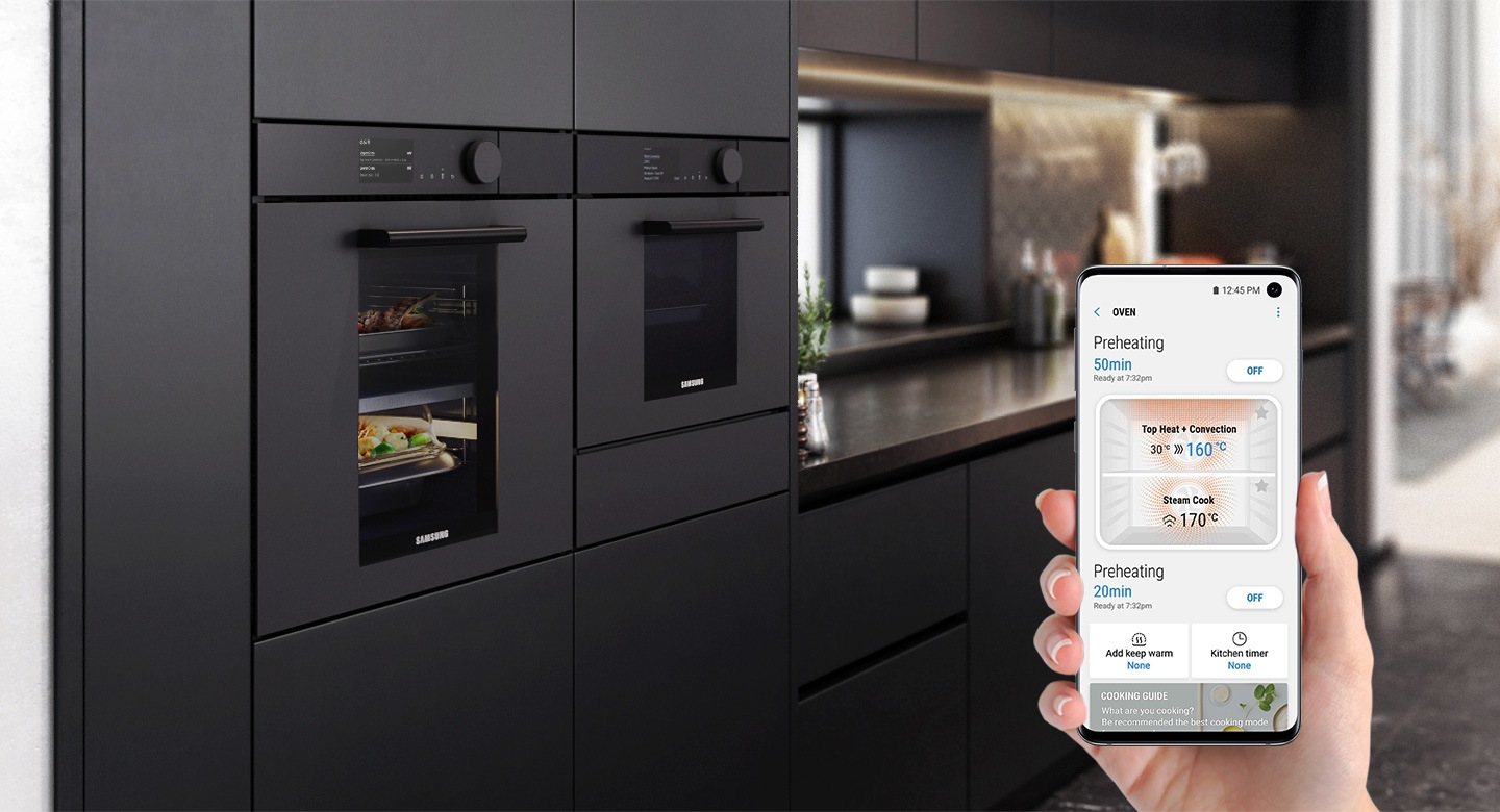 Scène de cuisine avec fours, un smartphone montrant les commandes de l'application et des meubles sombres.