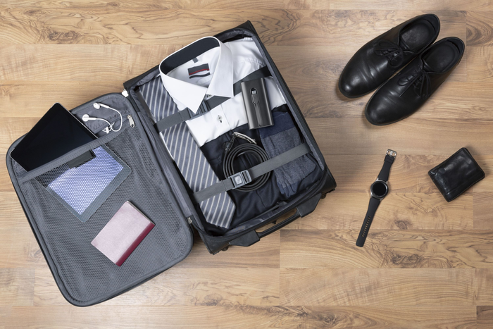 Offener Koffer auf Holzboden mit Kleidung, Schuhen, Uhr, Geldbörse und Tablet. Draufsicht.
