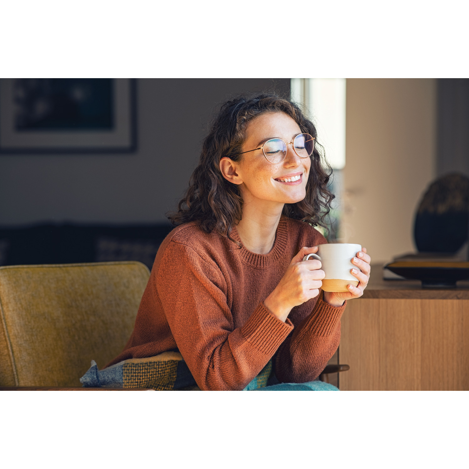 A woman holding a mug, smiling. Wearing glasses and a brown sweater.