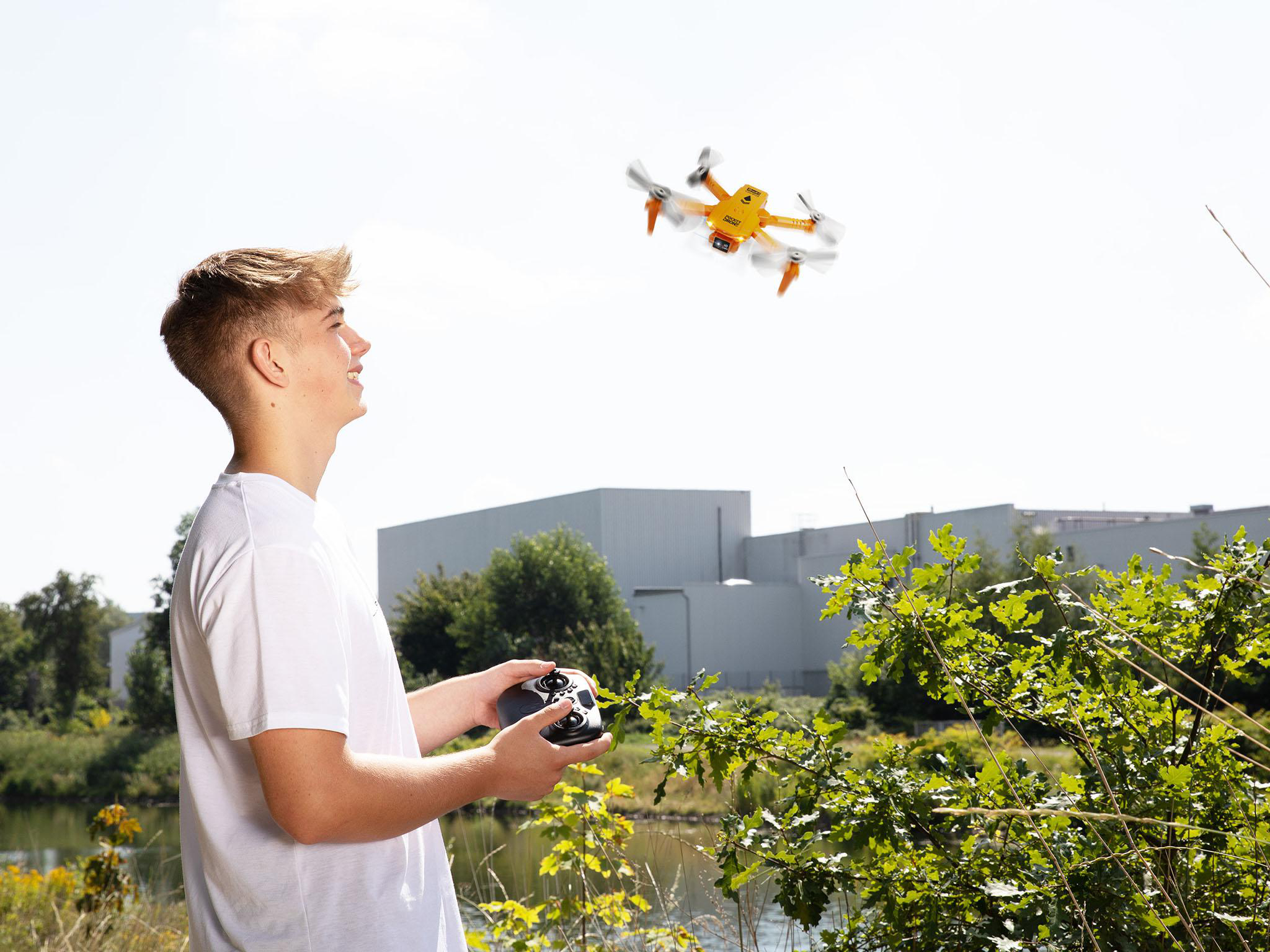 Ein junger Mann bedient eine Drohne. Eine gelbe Drohne fliegt. Bäume und ein Gebäude im Hintergrund.