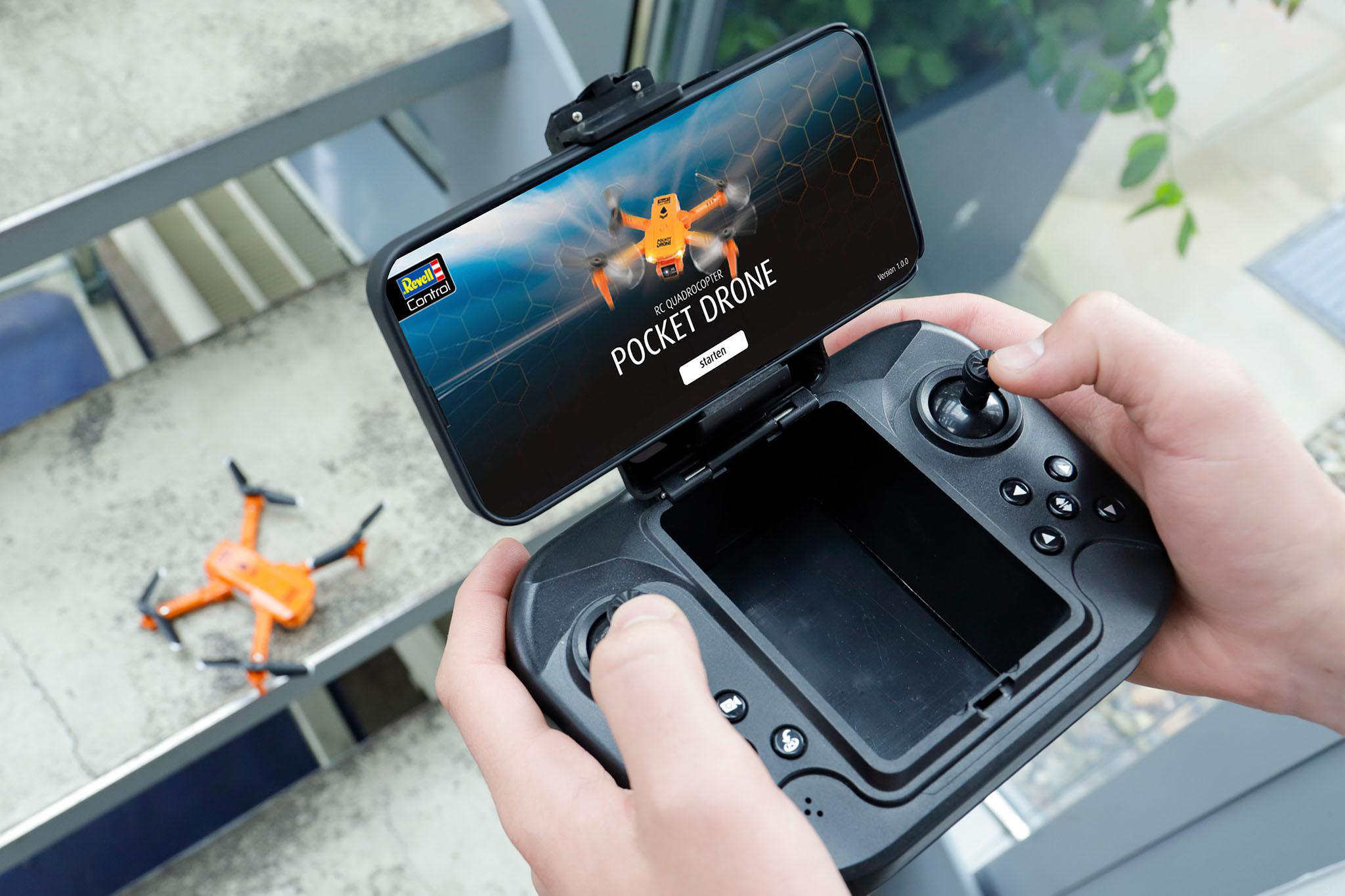 Hände halten eine Drohnen-Fernbedienung mit Telefon, das Drohnen anzeigt. Eine orangefarbene Drohne liegt daneben.