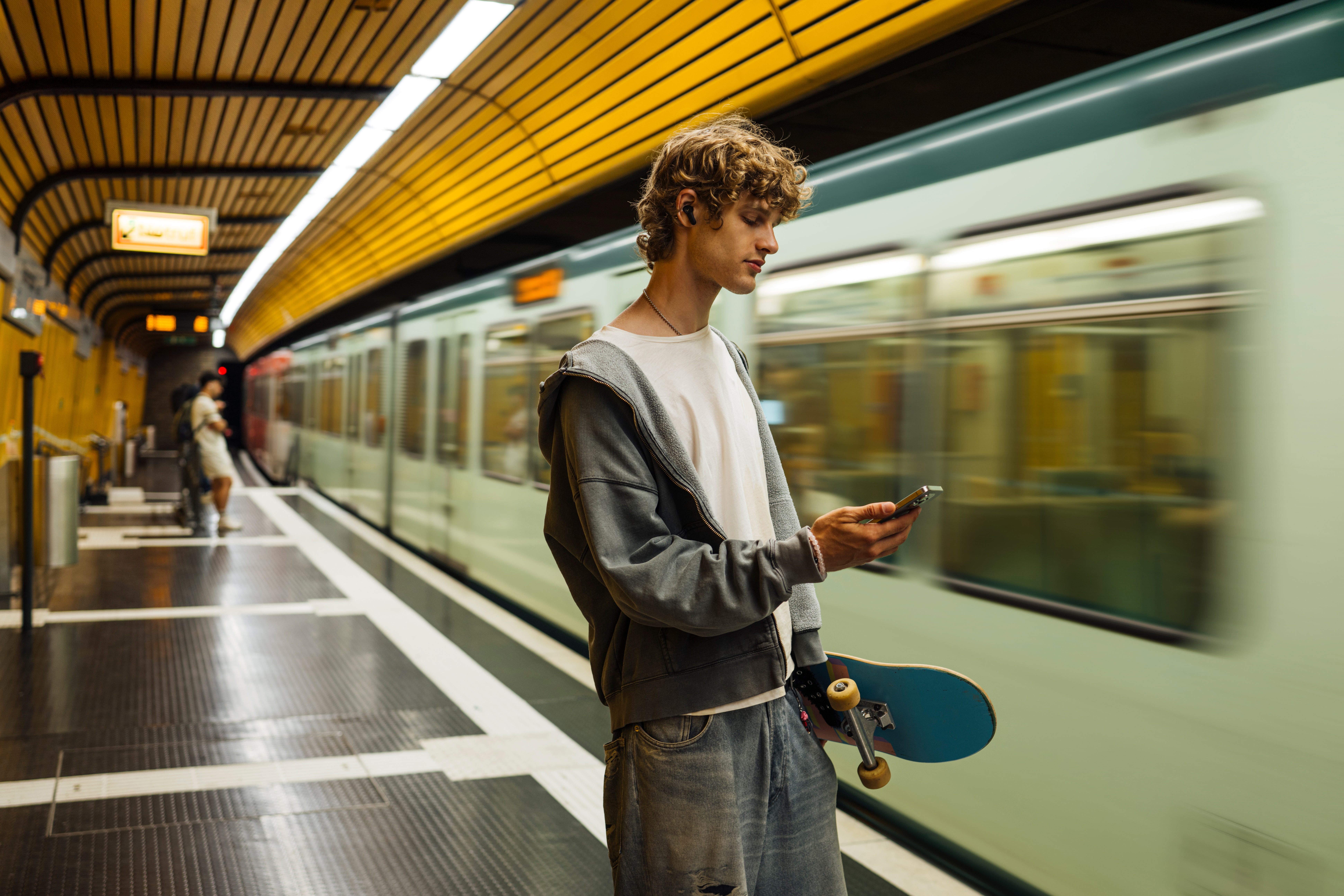 Ein junger Mann wartet in einer U-Bahn-Station, hält ein Skateboard und schaut auf sein Handy.