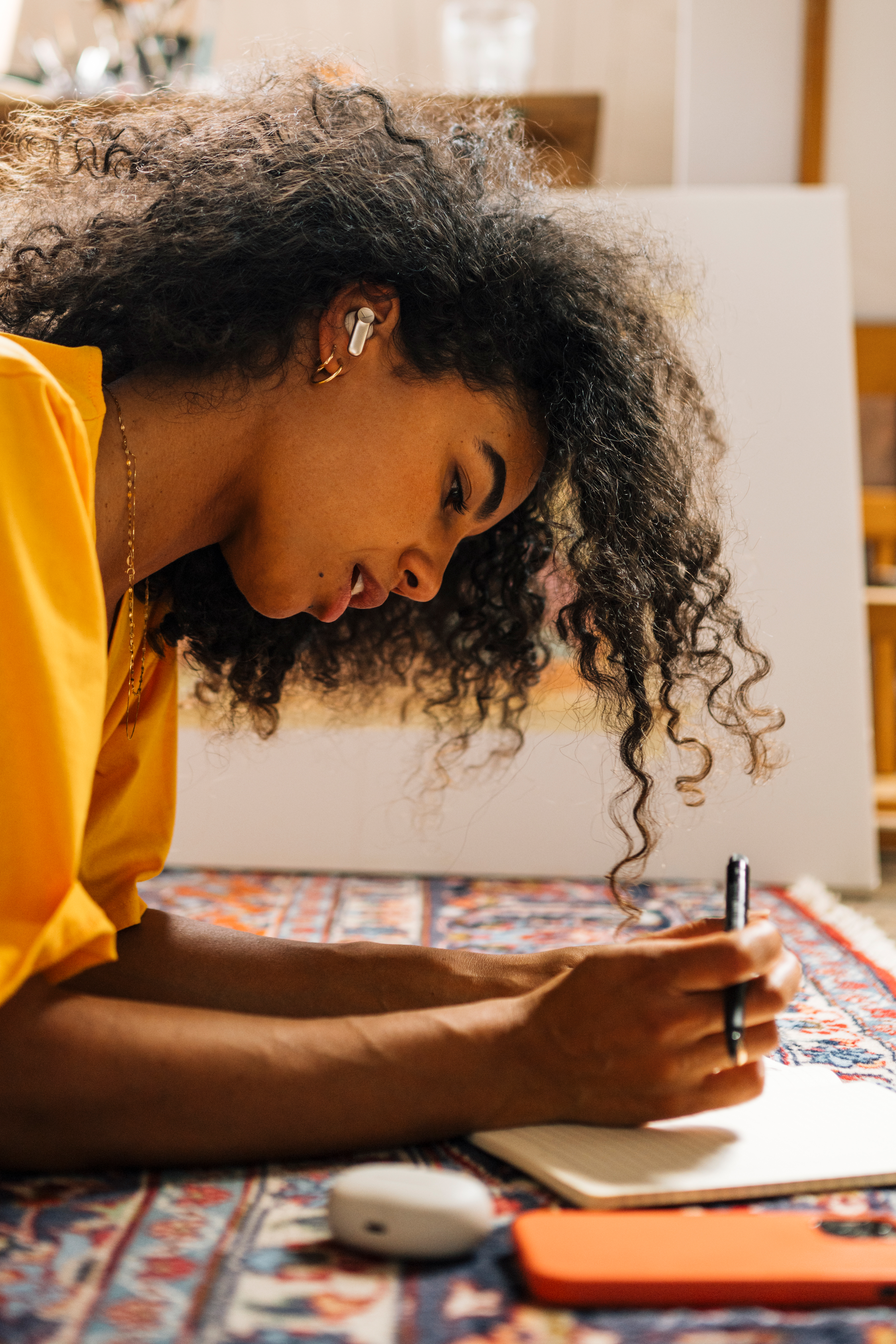Una donna con i capelli ricci scrive su un quaderno, sdraiata su un tappeto colorato, con penna e auricolari.