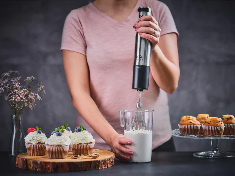 Egy rózsaszín inget viselő személy kézi mixerrel habot ver. Cupcake-ek fa deszkán.
