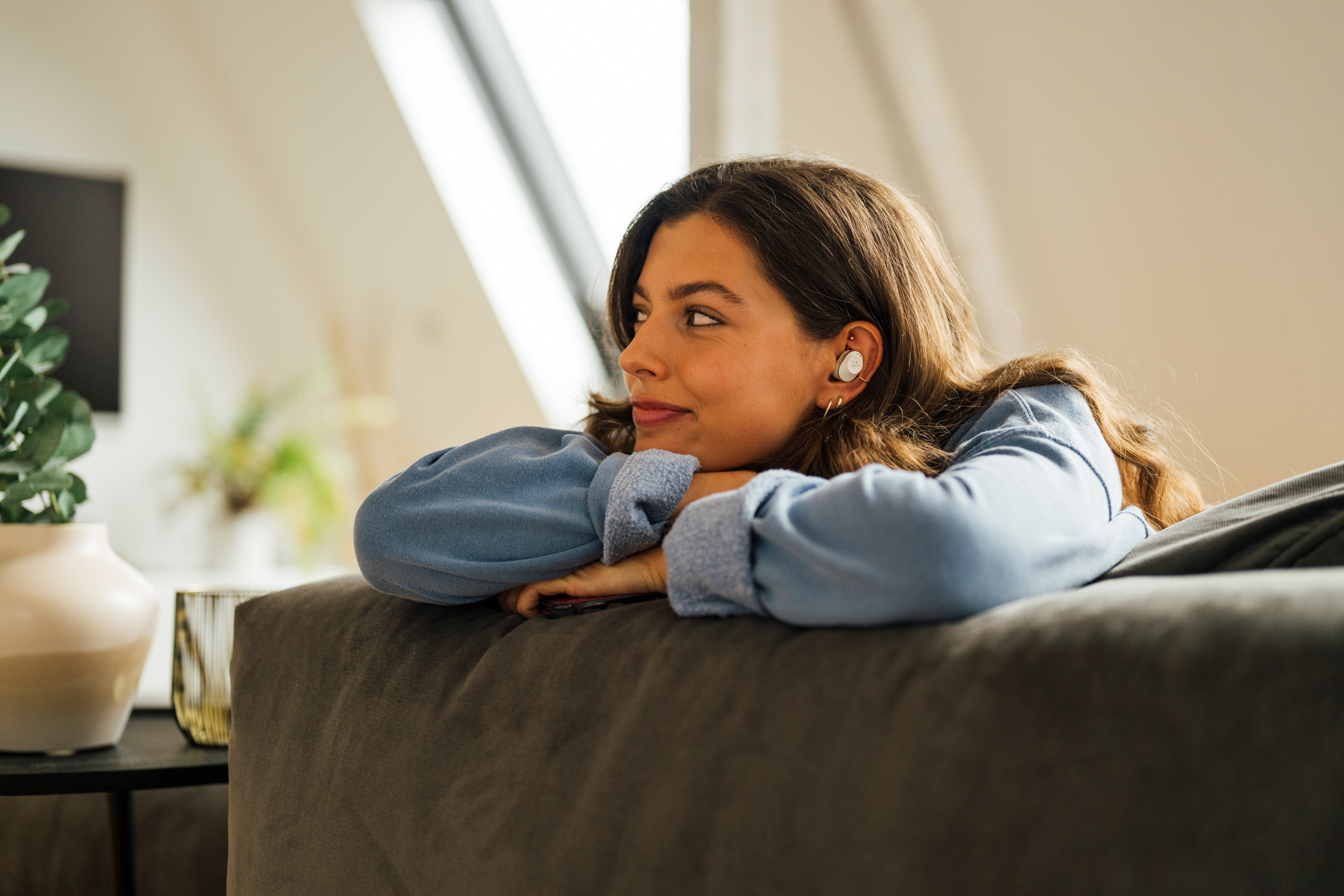Eine Frau lehnt sich auf einem Sofa und trägt ein blaues Hemd. Sie schaut auf etwas außerhalb des Bildes.