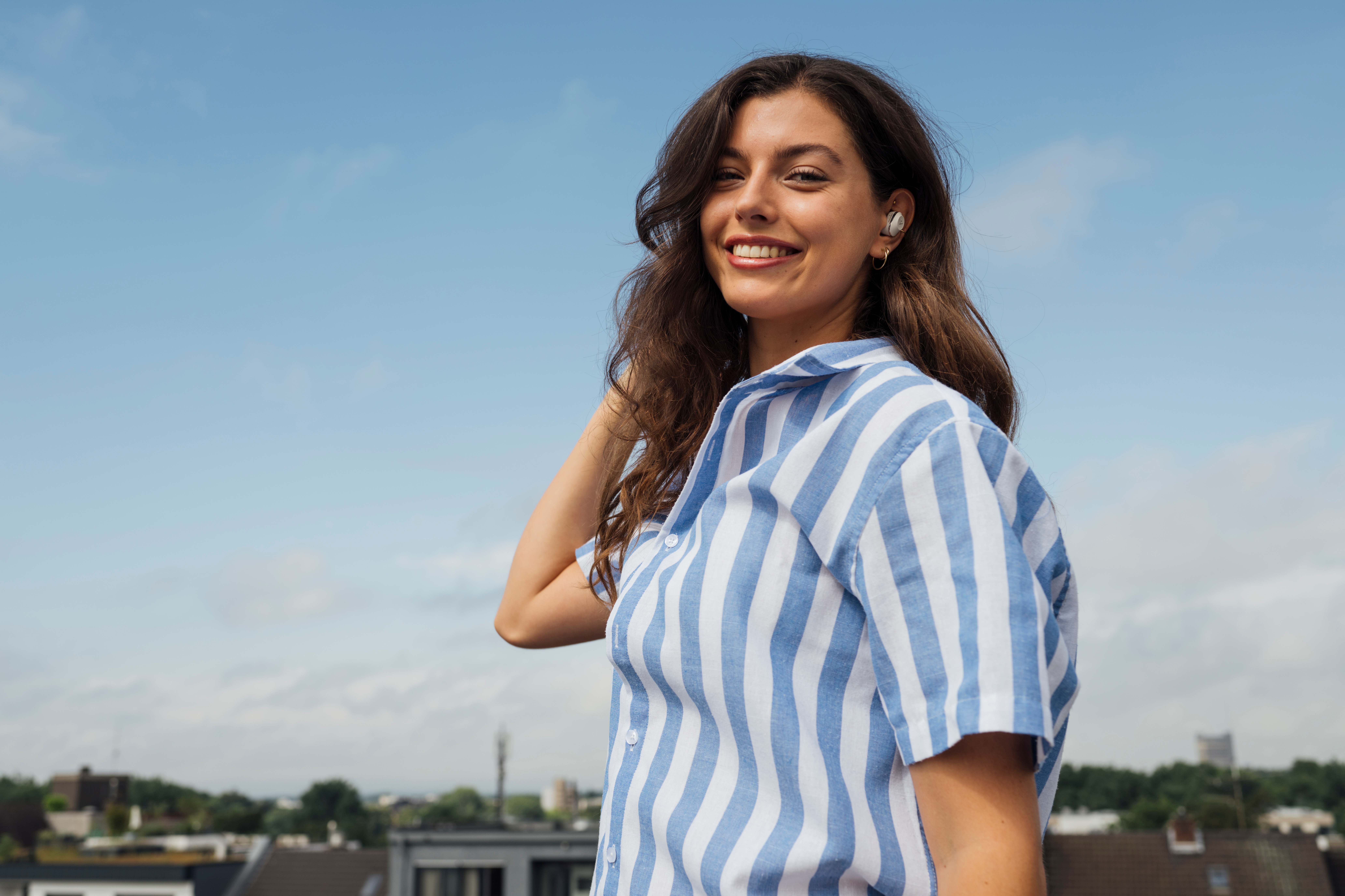 Eine Frau mit langen Haaren, die Ohrhörer trägt, lächelt. Sie ist auf einem Dach mit blauem Himmel.