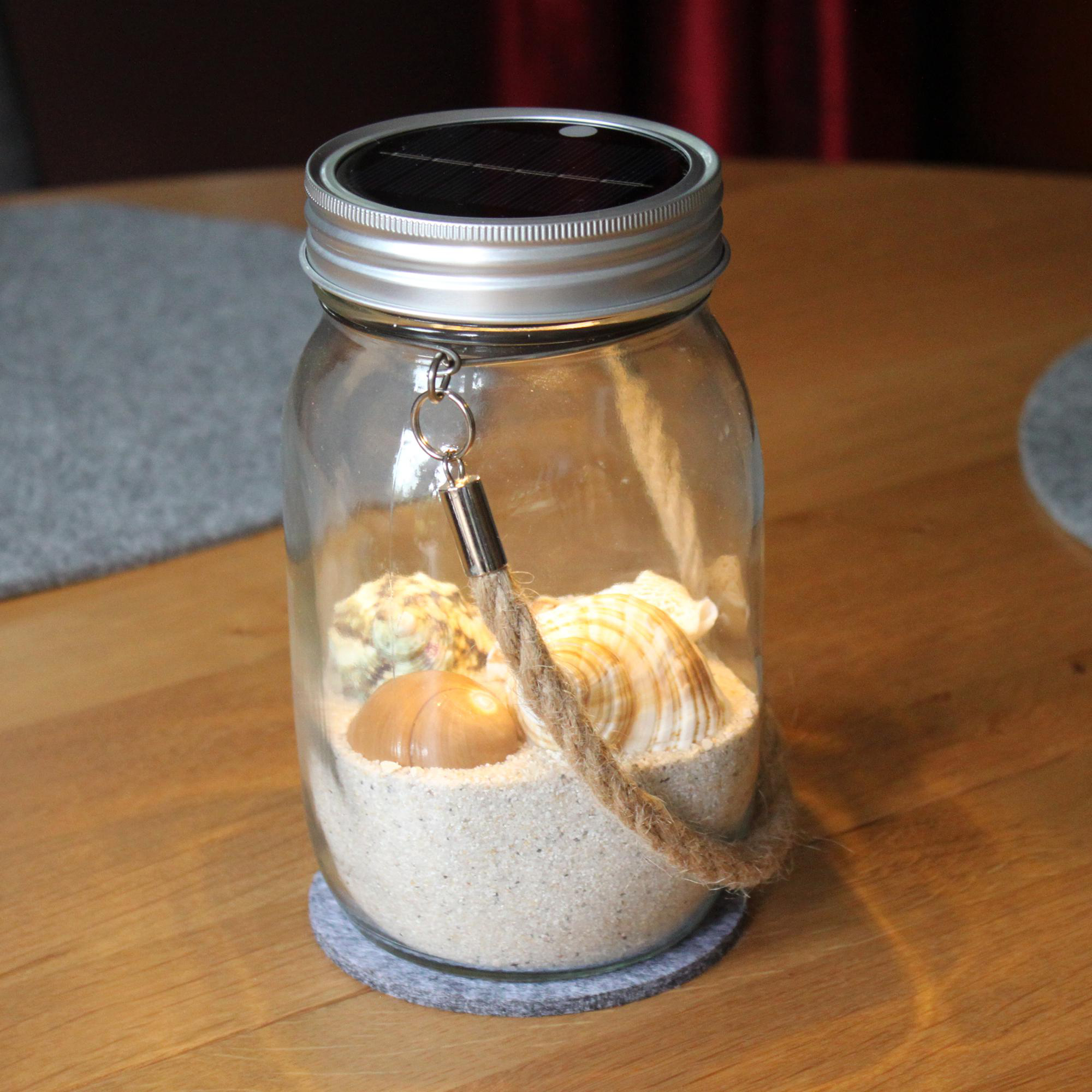 Glasbehälter mit Sand, Muscheln und Seilgriff, mit Solarpanel.