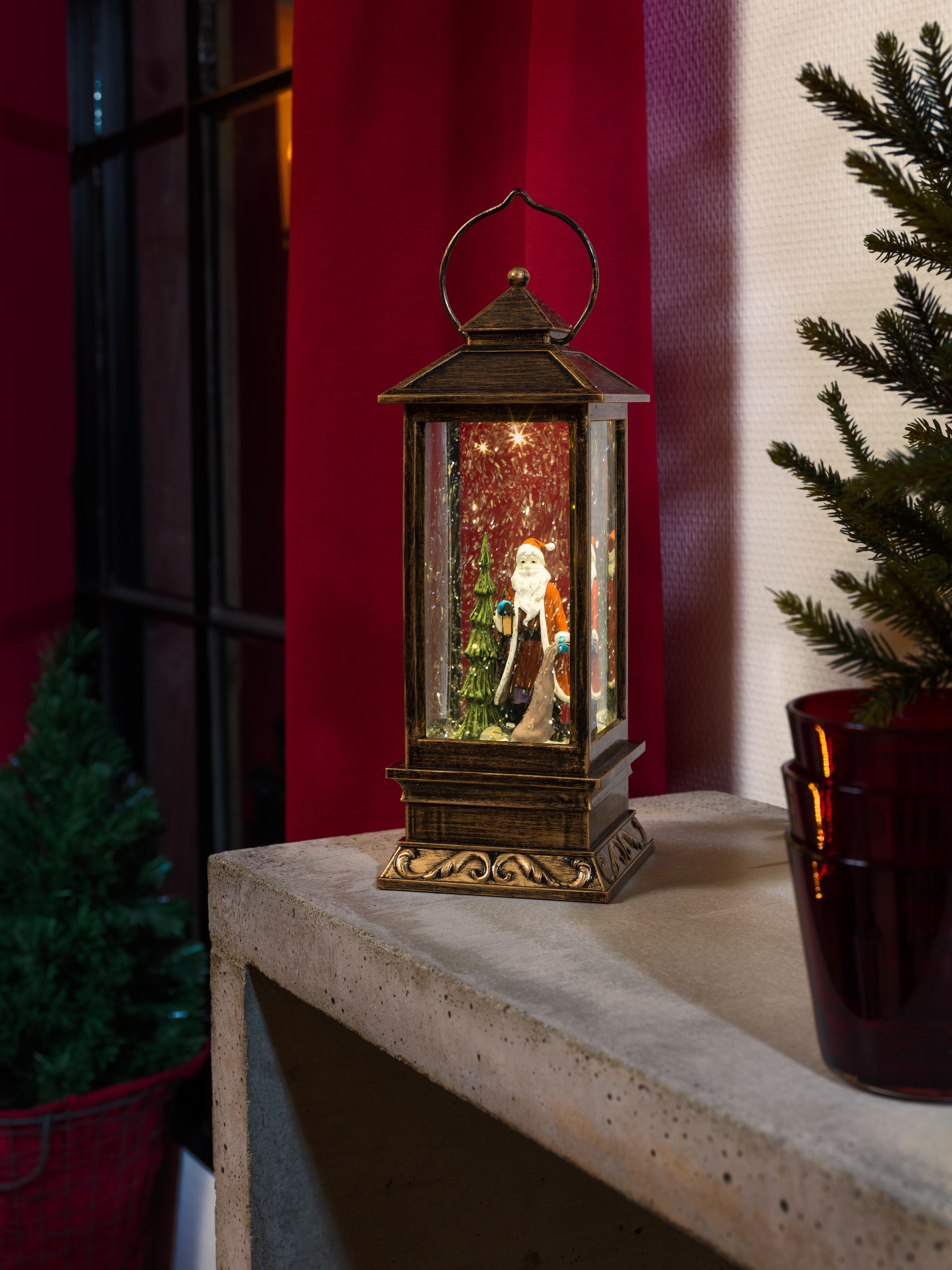 Eine beleuchtete Laterne mit Weihnachtsmannfigur auf einer Steinoberfläche, roter Wand und Baum im Hintergrund.