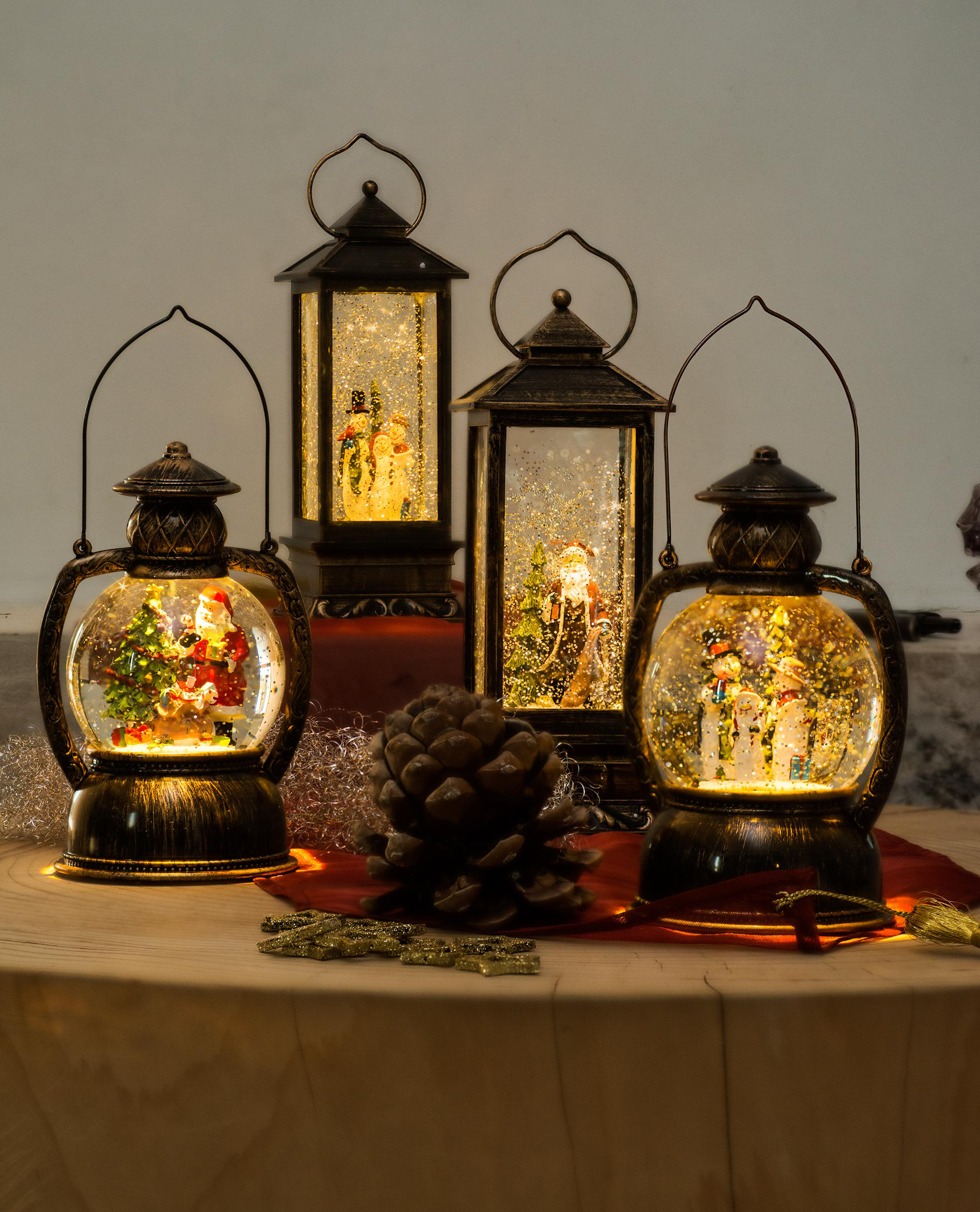 Vier Weihnachtslaternen auf einem Holztisch mit Ornamenten. Jede hat eine Schneeszene mit Figuren.