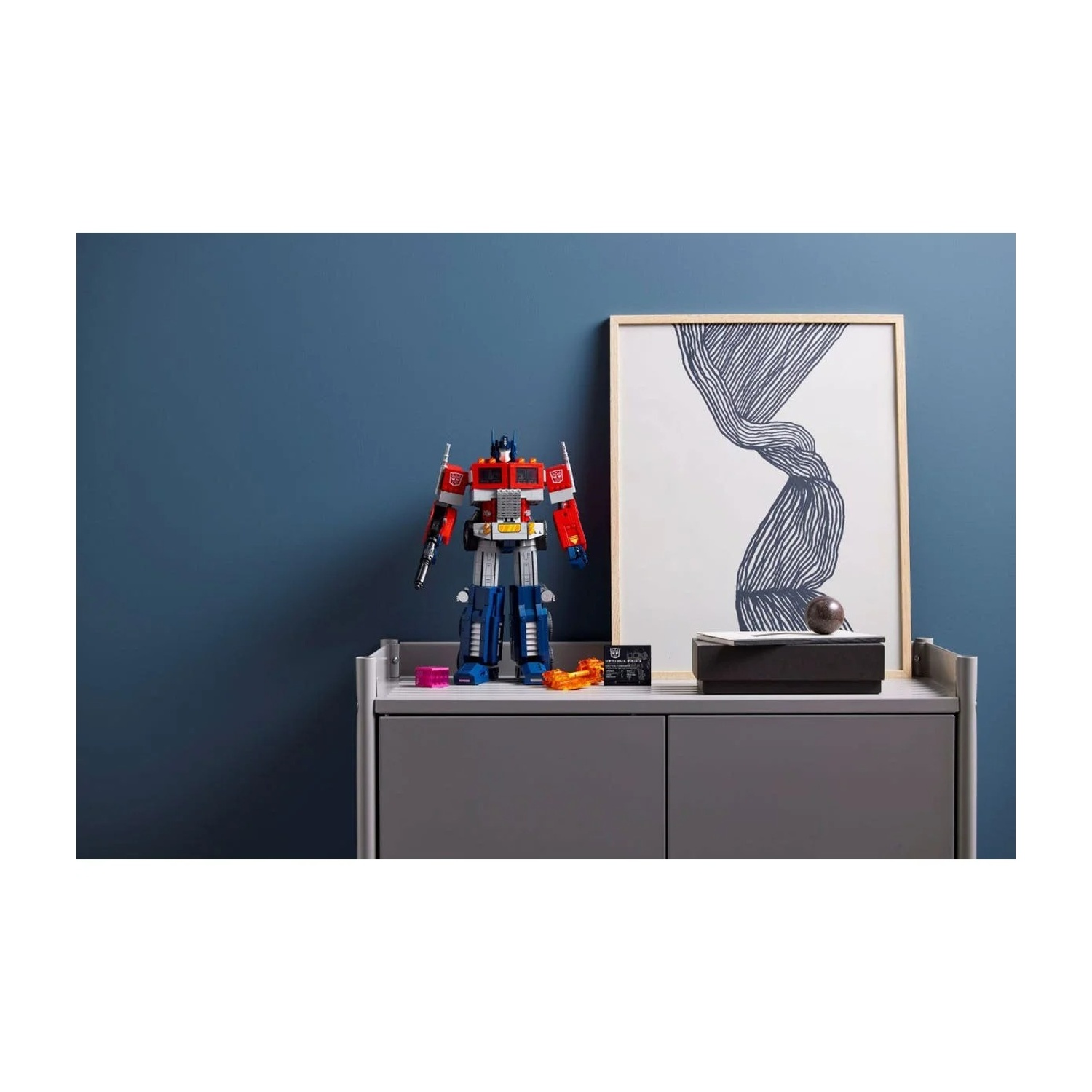 Un giocattolo robot Lego Optimus Prime con un dipinto e altri oggetti su una mensola grigia contro un muro blu.