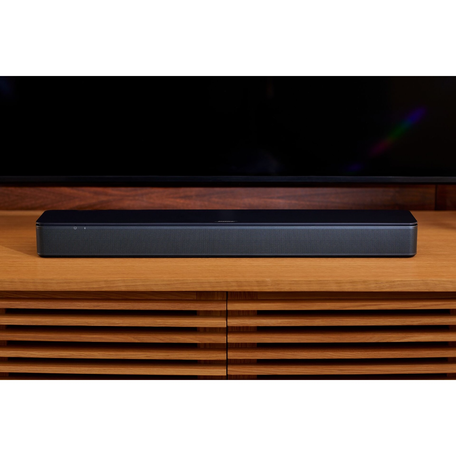 Una soundbar nera su un mobile in legno. Una TV nera sullo sfondo.