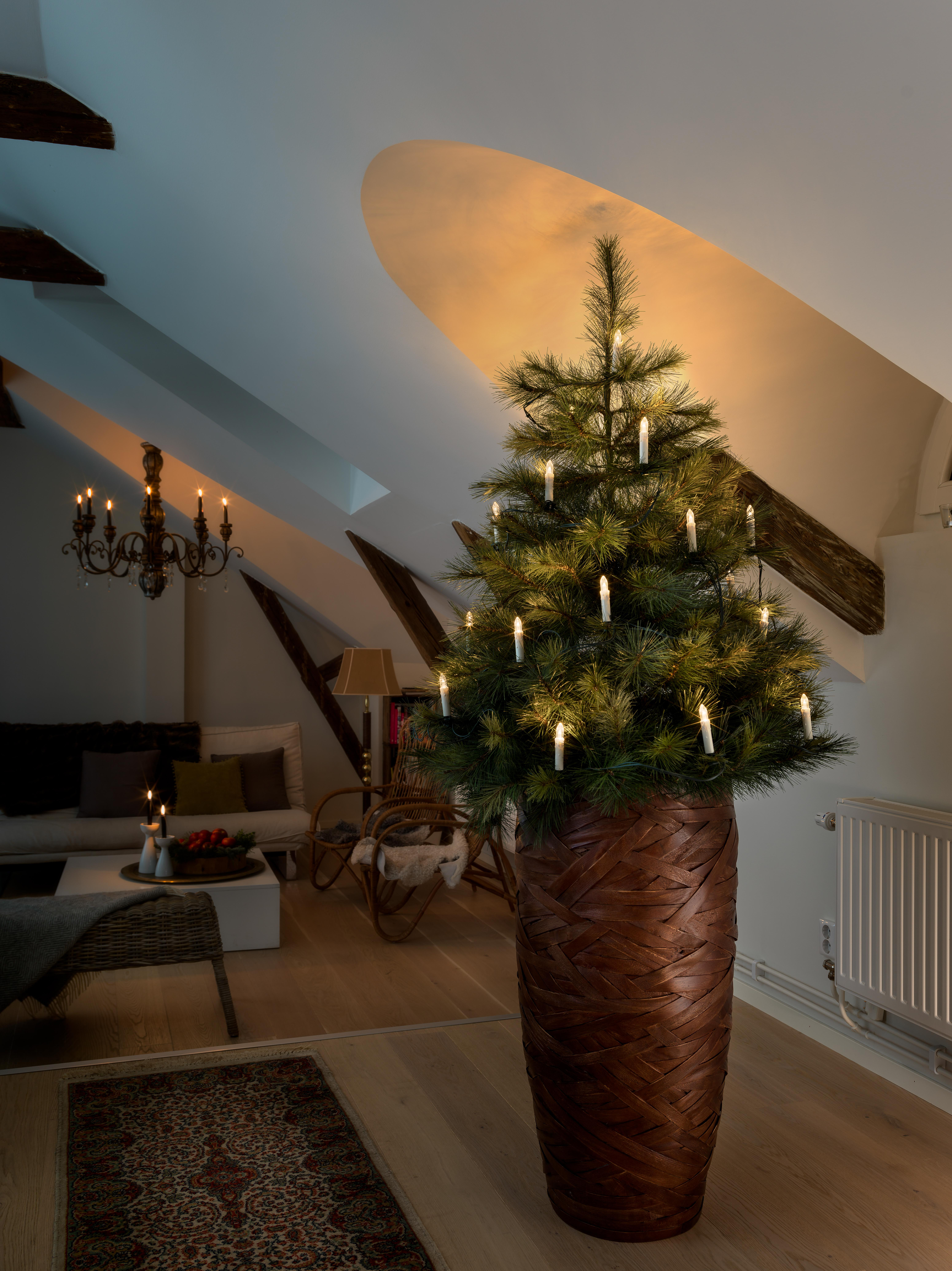 Ein beleuchteter Weihnachtsbaum in einem hohen braunen Topf in einem Raum mit hoher Decke.