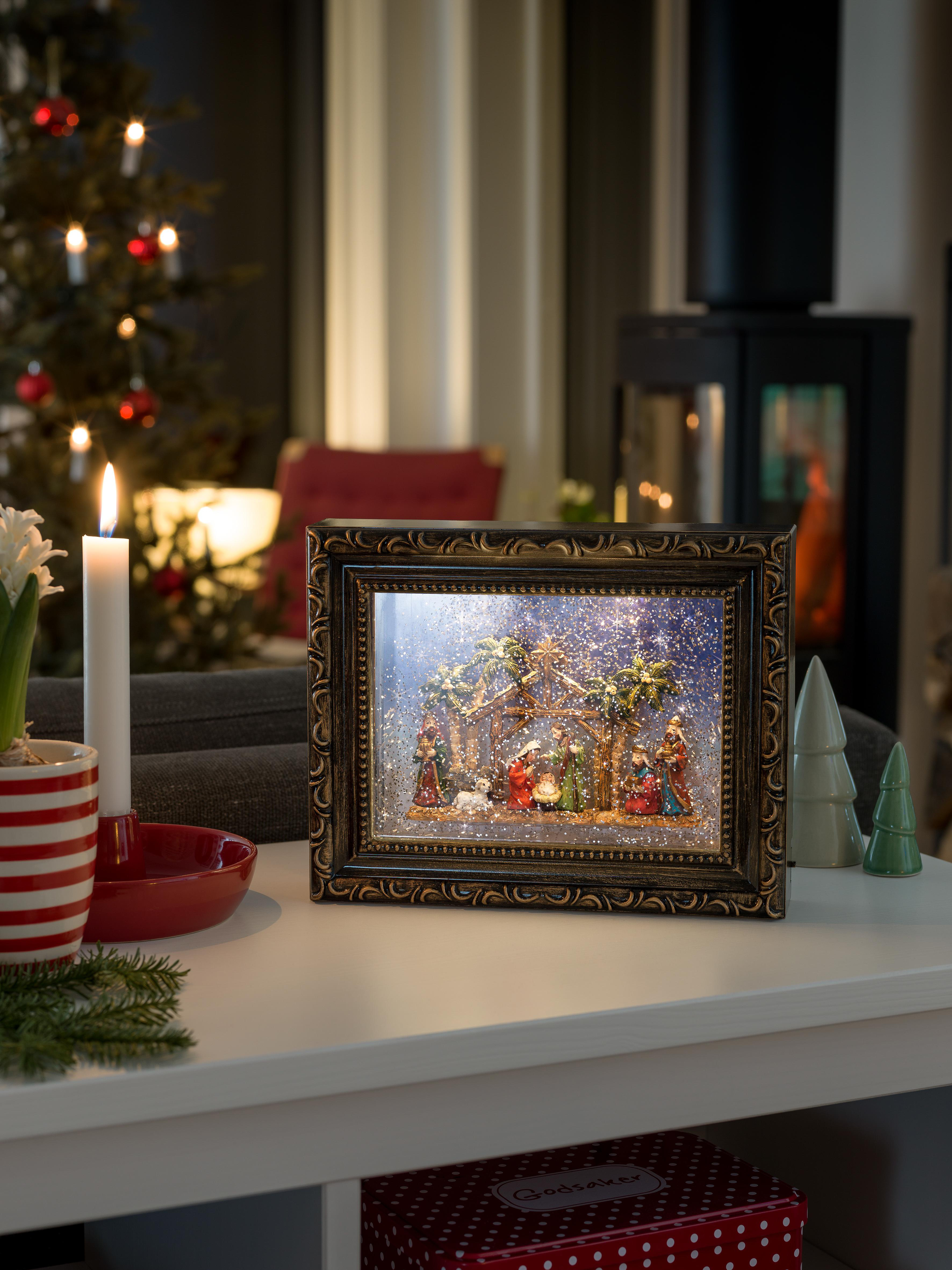 Weihnachtsszene in einem Rahmen mit Krippe auf einem Tisch, brennender Kerze und Weihnachtsbaum.