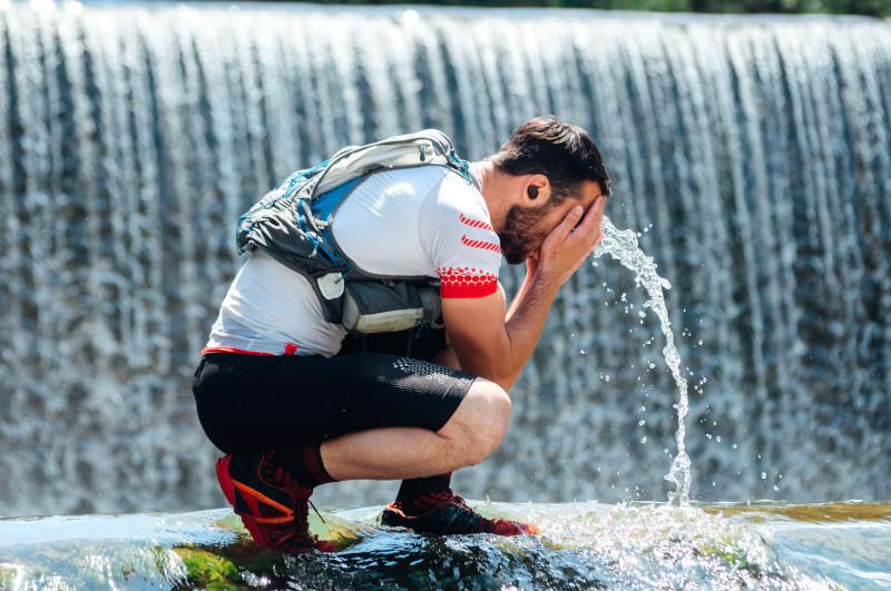 Ein Mann hockt sich in der Nähe eines Wasserfalls und spritzt sich Wasser ins Gesicht.