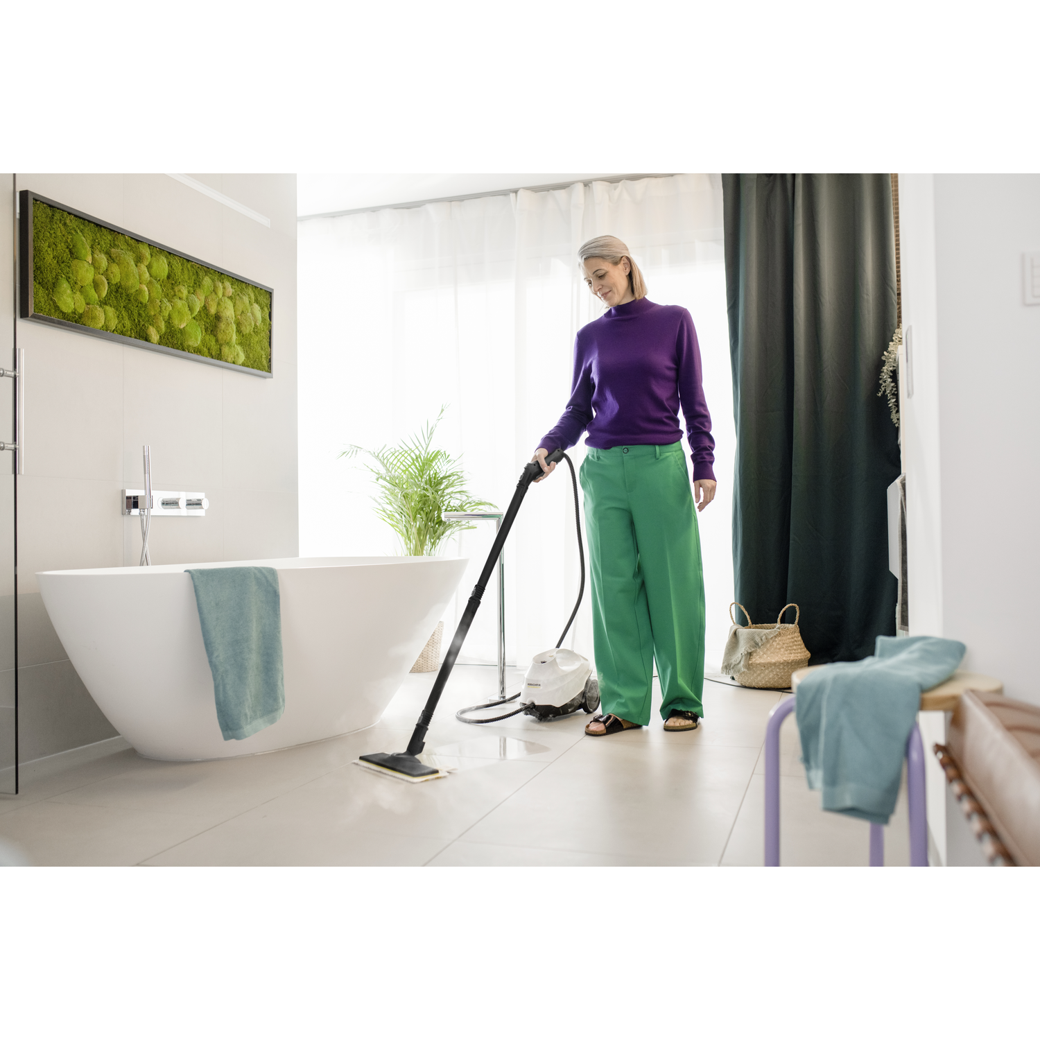 Woman in green pants cleaning bathroom with steam cleaner; white tub, plant, and towels.