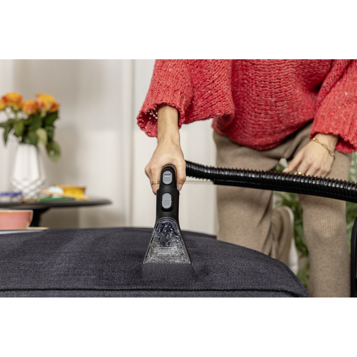 Person cleaning a dark gray cushion with a vacuum cleaner. A red sweater is visible. Background: flowers in a vase.