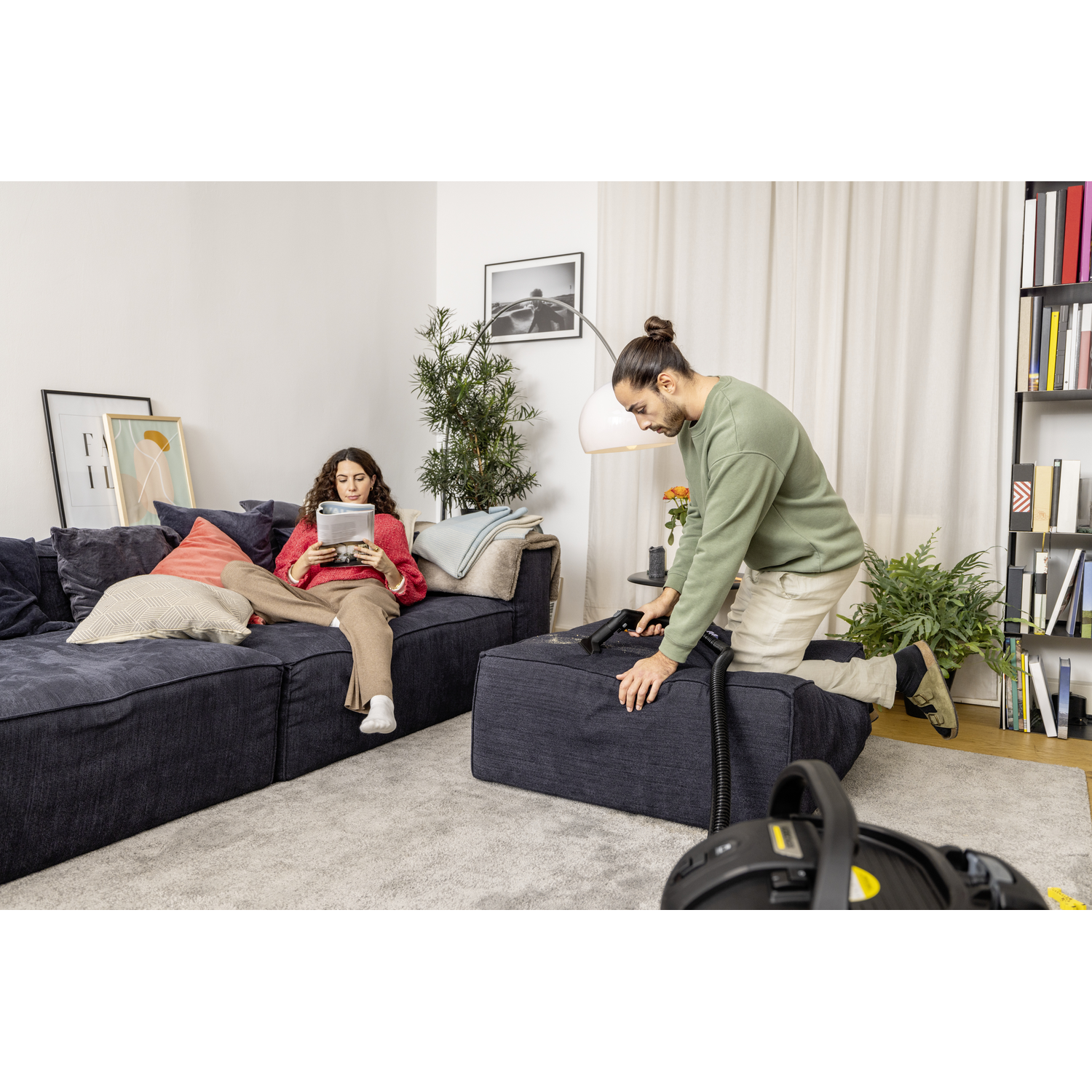 Man vacuuming a dark sofa in a living room. Woman on sofa uses a tablet. Carpet on floor.