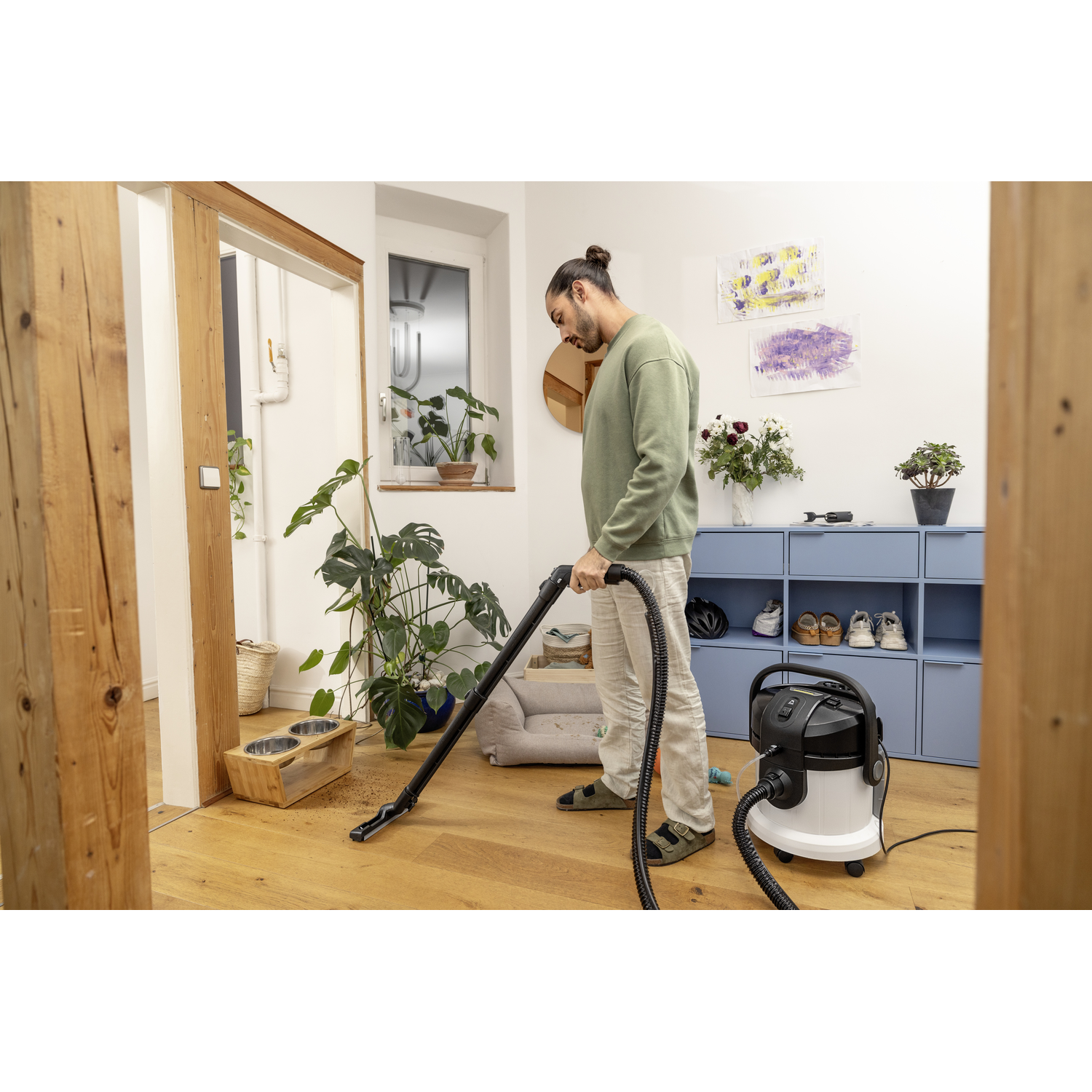 Man vacuuming a wooden floor with a vacuum cleaner, in a room with furniture and plants.