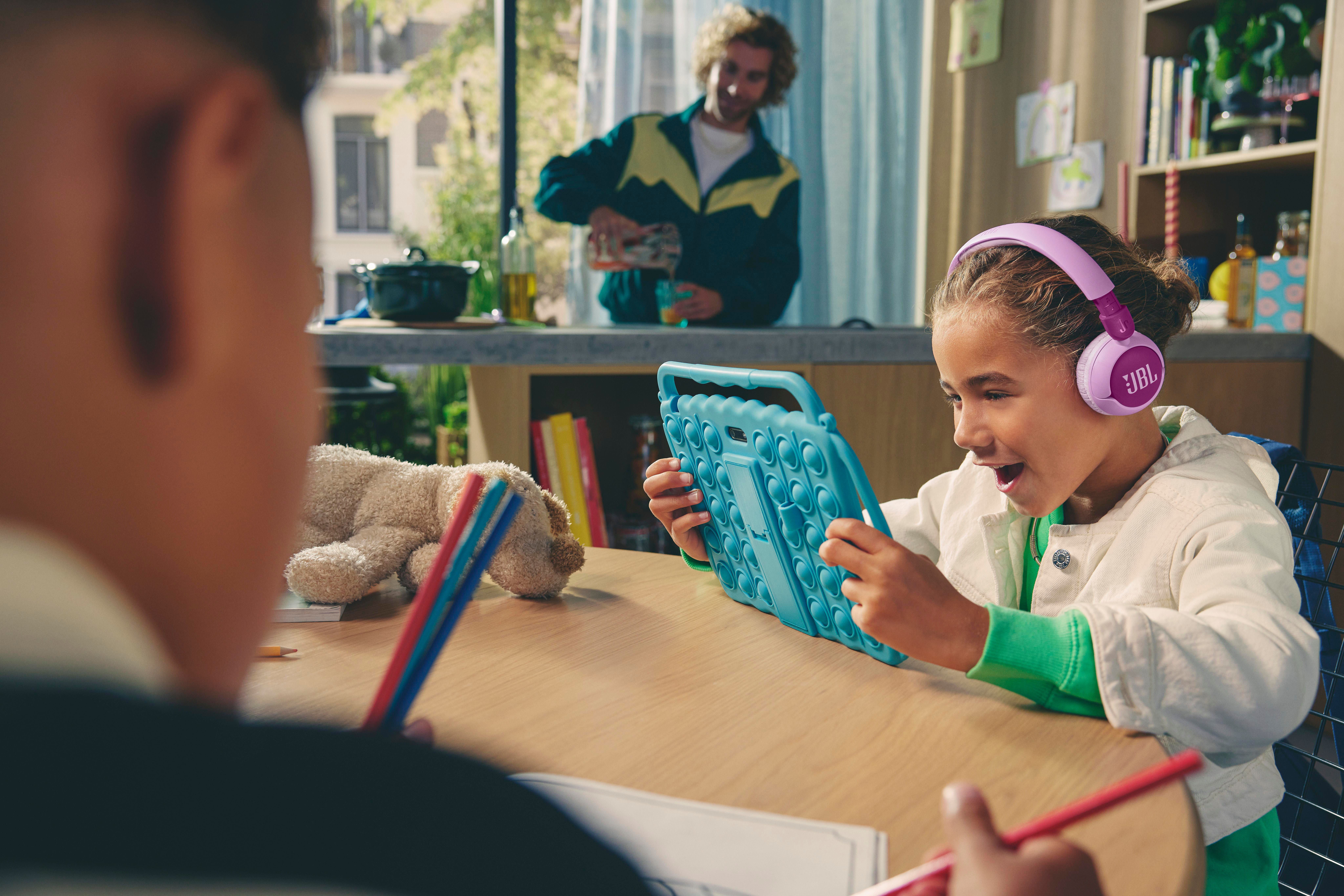 Ein Mädchen mit Kopfhörern benutzt ein Tablet, mit Mann und Objekten im Hintergrund.