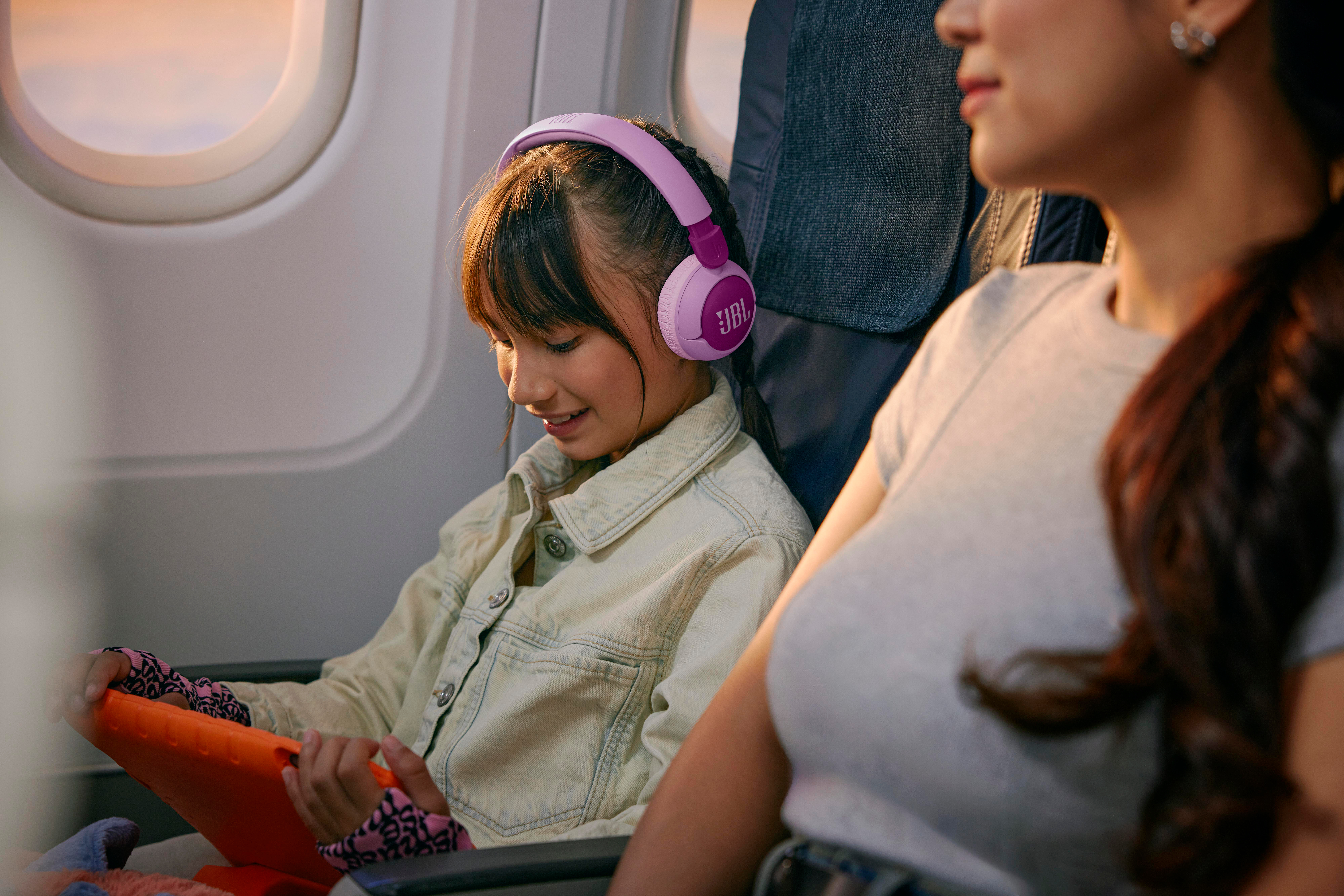 Ein Mädchen mit lila Kopfhörern in einem Flugzeug. Ihre Mutter ist teilweise sichtbar.