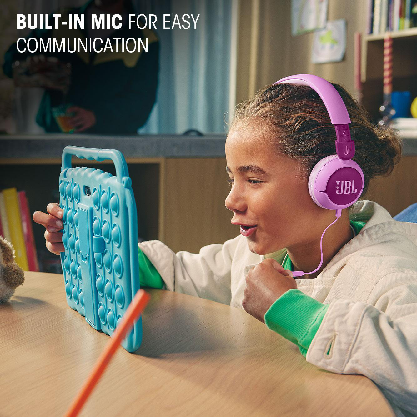 Mädchen mit lila JBL-Kopfhörern schaut auf ein blaues Pop-it-Tablet. Integriertes Mikrofon für einfache Kommunikation.