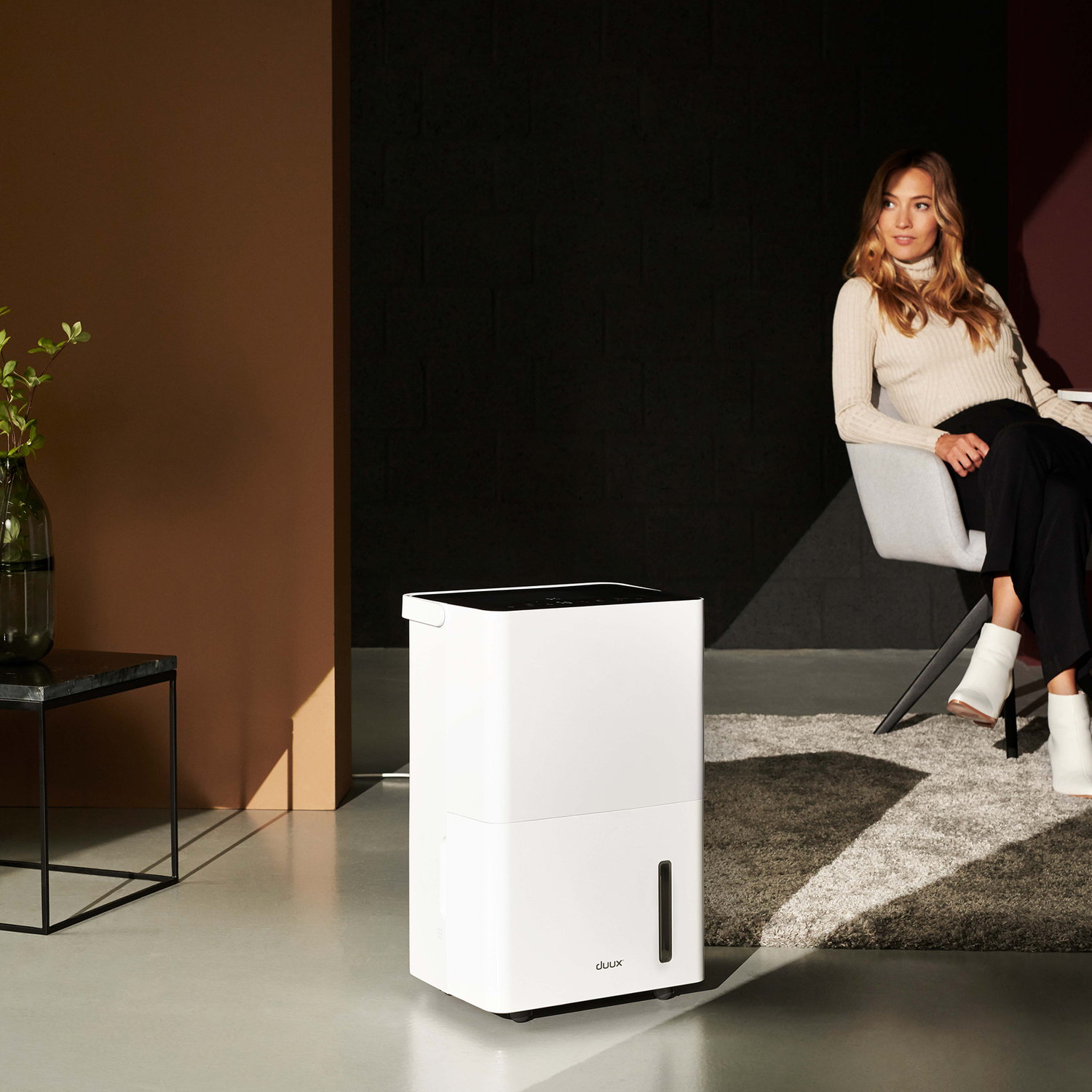 Vrouw zit bij een witte luchtontvochtiger en een kleine tafel met plant.