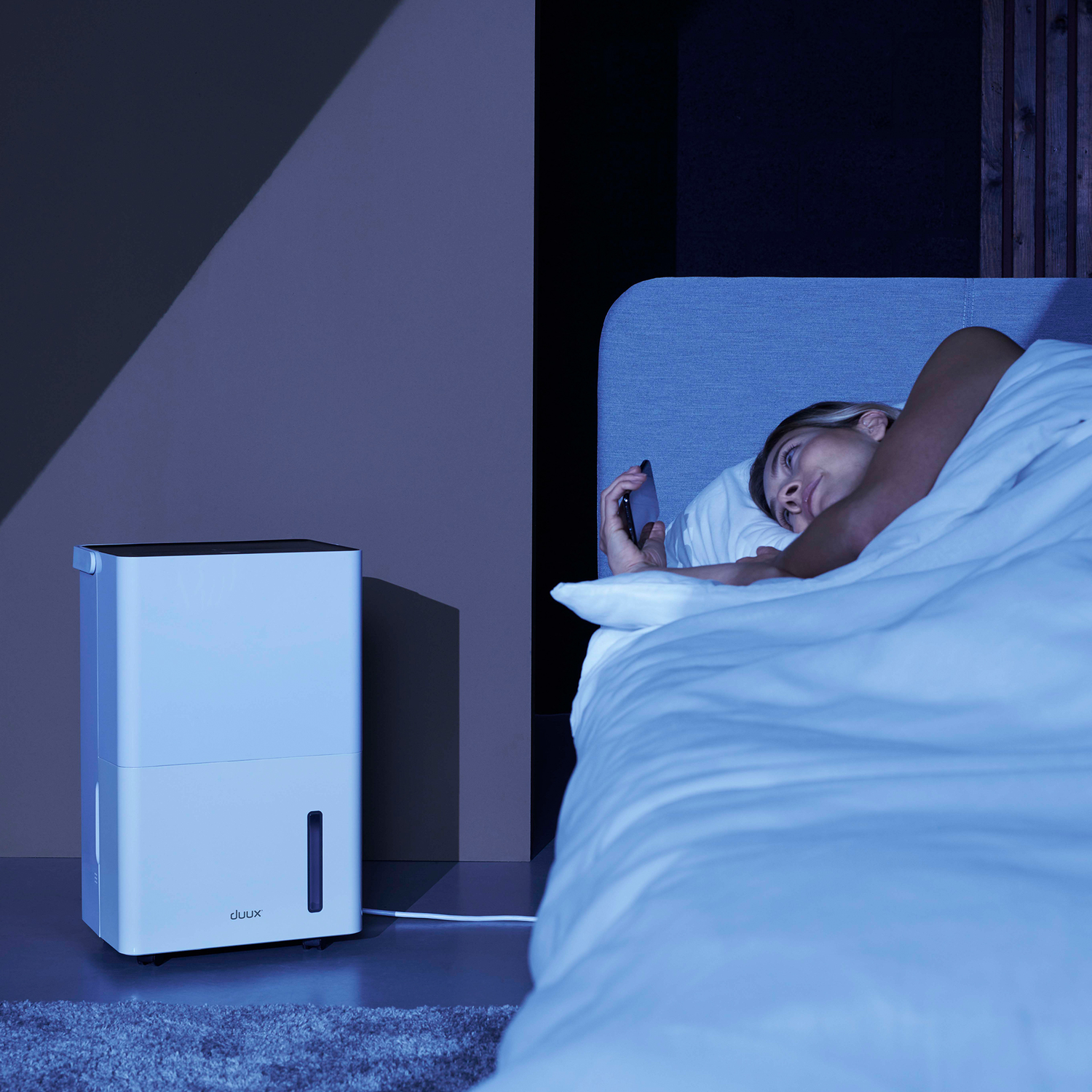 Een vrouw in bed gebruikt haar telefoon in de buurt van een witte luchtontvochtiger in een zwak verlichte kamer.