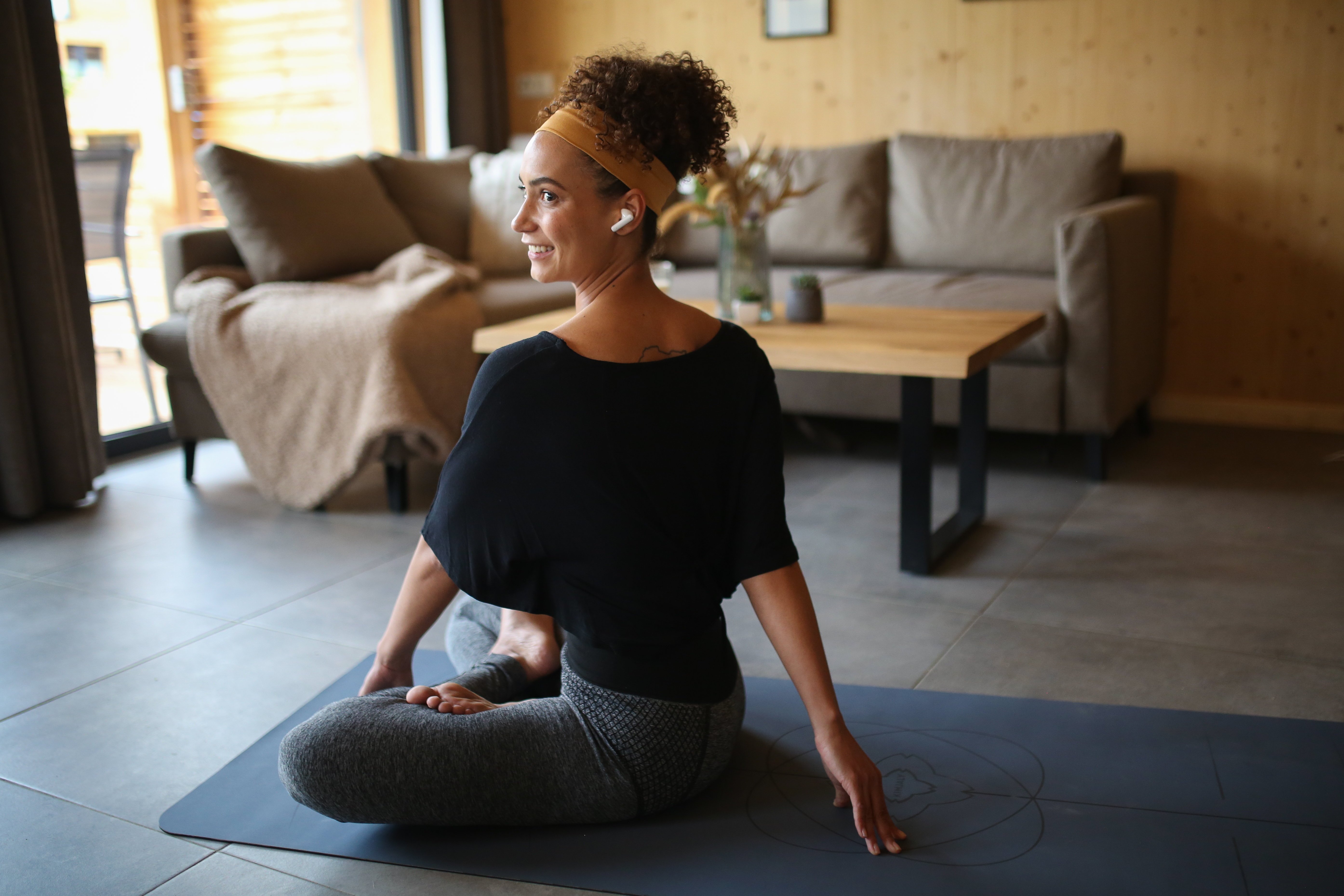 Eine Frau macht Yoga auf einer Matte, trägt Ohrhörer. Innenraum, mit Sofa und Tisch.