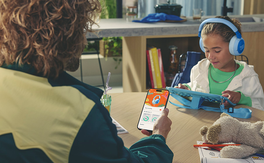 Eine Person hält ein Telefon mit geöffneter App. Ein Kind trägt Kopfhörer und schaut auf ein Tablet.