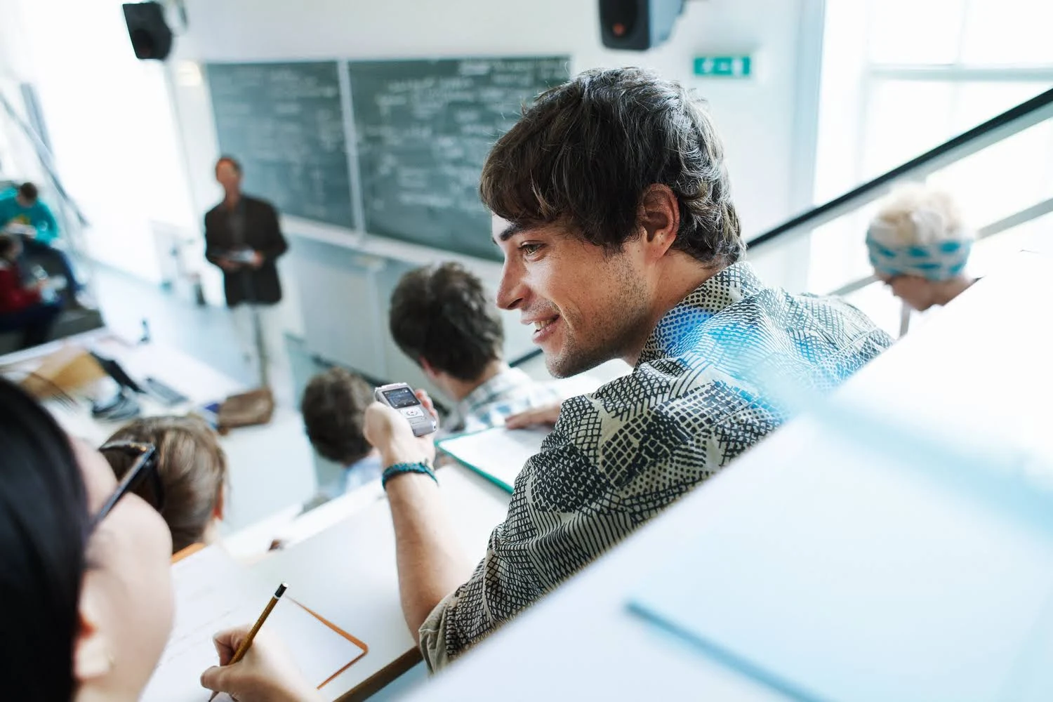 Un estudiante en un aula mira su teléfono. Se ven otros estudiantes y un profesor.