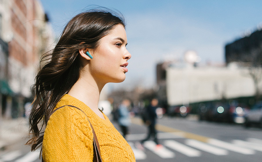 Una donna con un maglione giallo che indossa un auricolare blu in una strada cittadina.