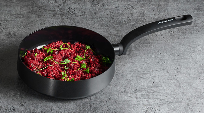 Czarna patelnia z ugotowanym czerwonym risotto i ziołami. Znajduje się na szarej powierzchni.