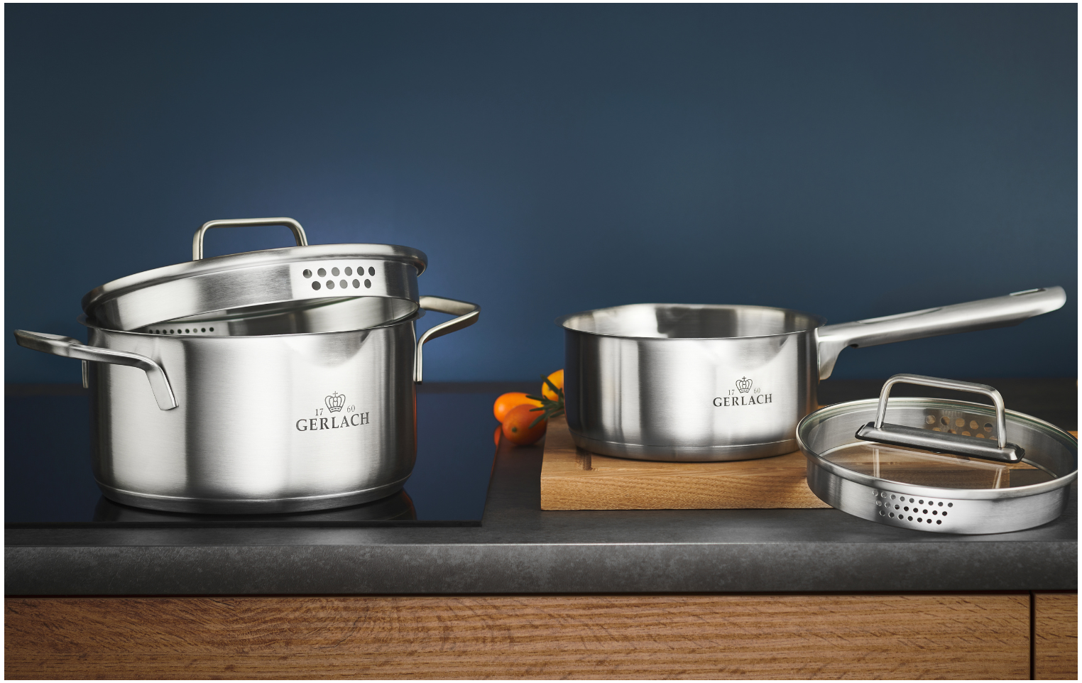 Garnki ze stali nierdzewnej i patelnia na czarnej płycie kuchennej. Obecna jest drewniana deska do krojenia.