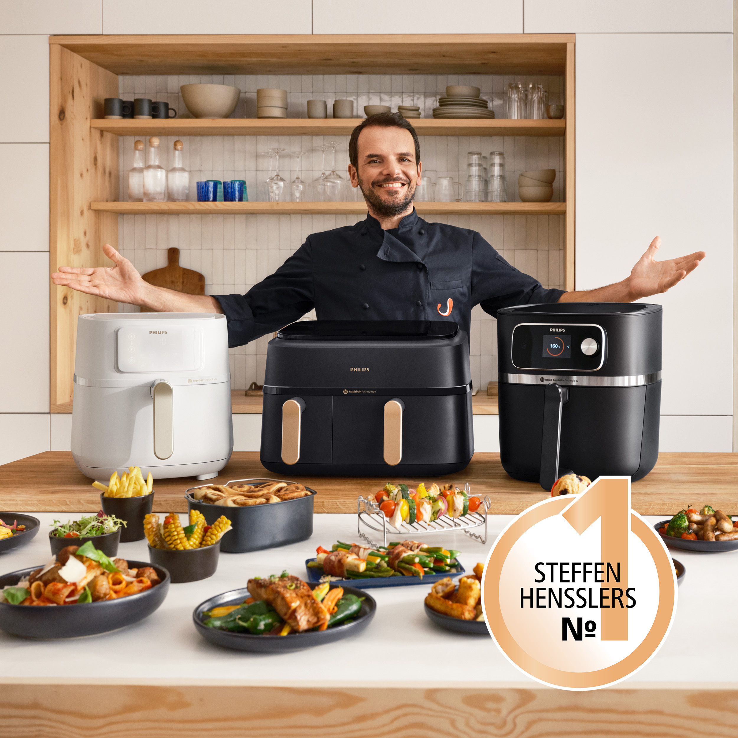 Homme en uniforme de chef présente des friteuses à air et des plats cuisinés sur une table en bois.