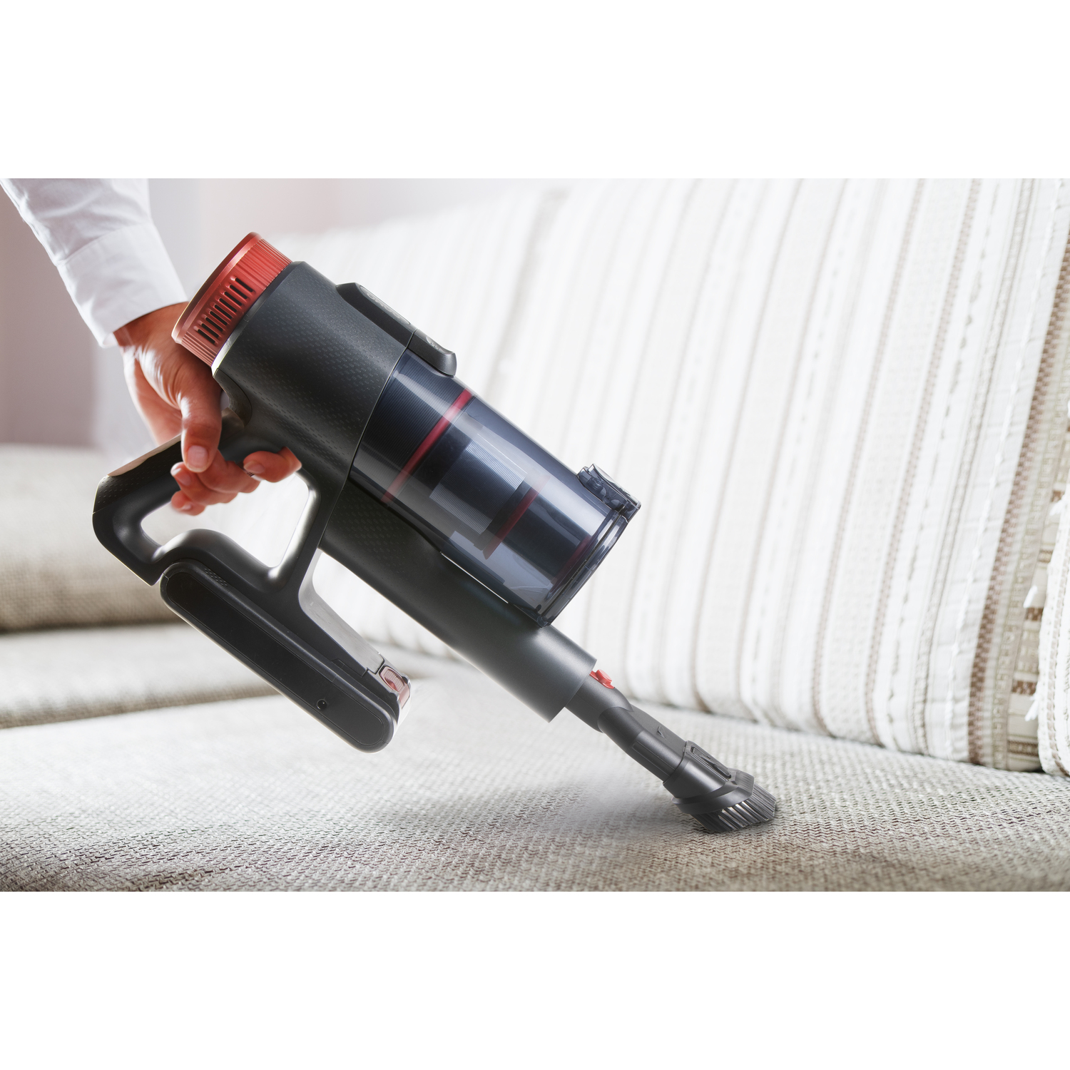 A handheld vacuum cleaner, black and red, is cleaning a beige sofa. A hand holds it.