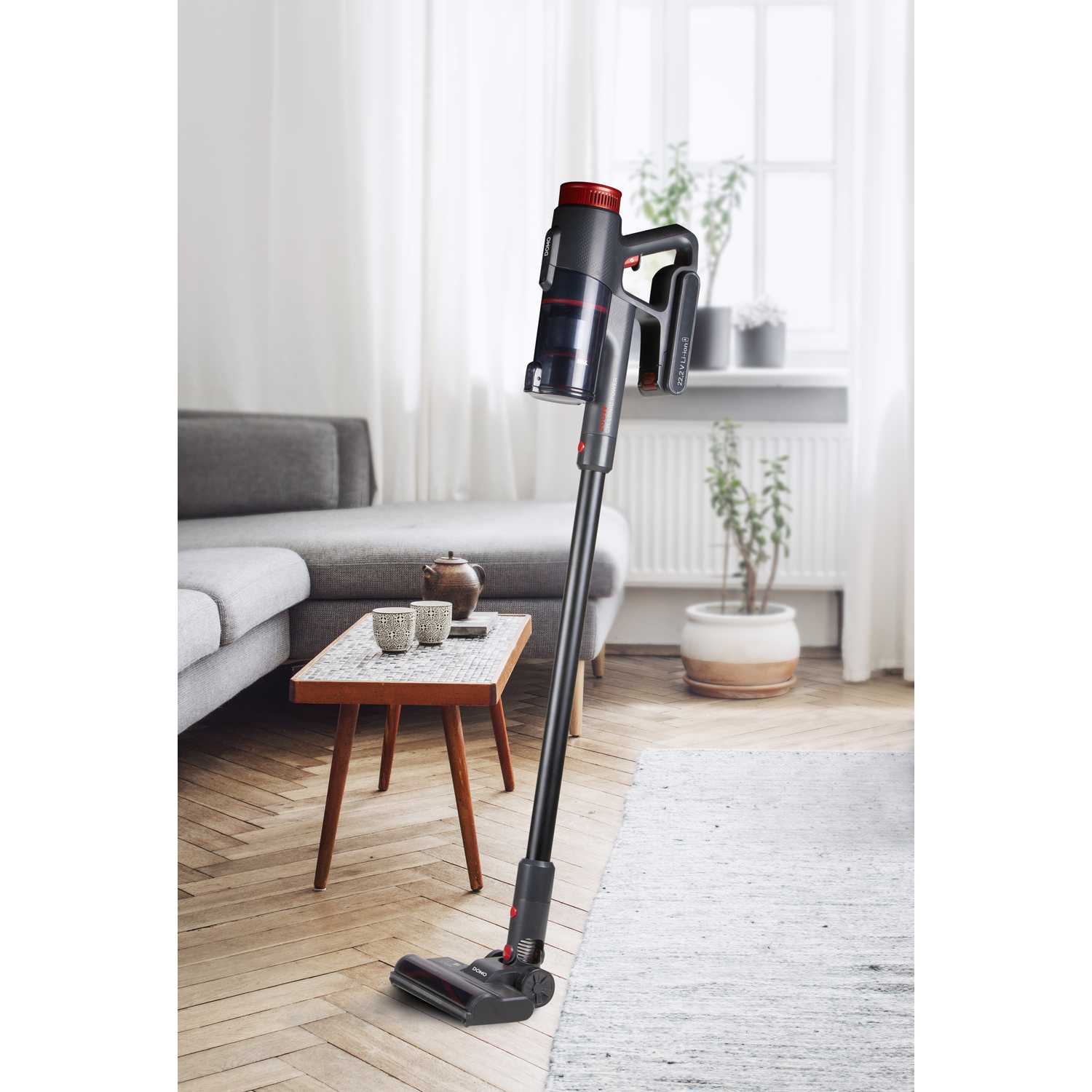 A black and red cordless vacuum cleaner stands in a living room, near a grey sofa and a wooden table.