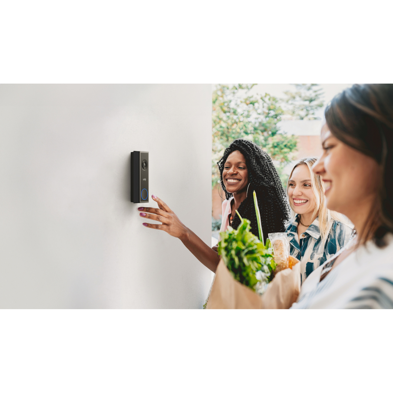 Trois femmes se tiennent près d'un mur blanc avec une sonnette. L'une montre, deux sourient, tenant des courses.