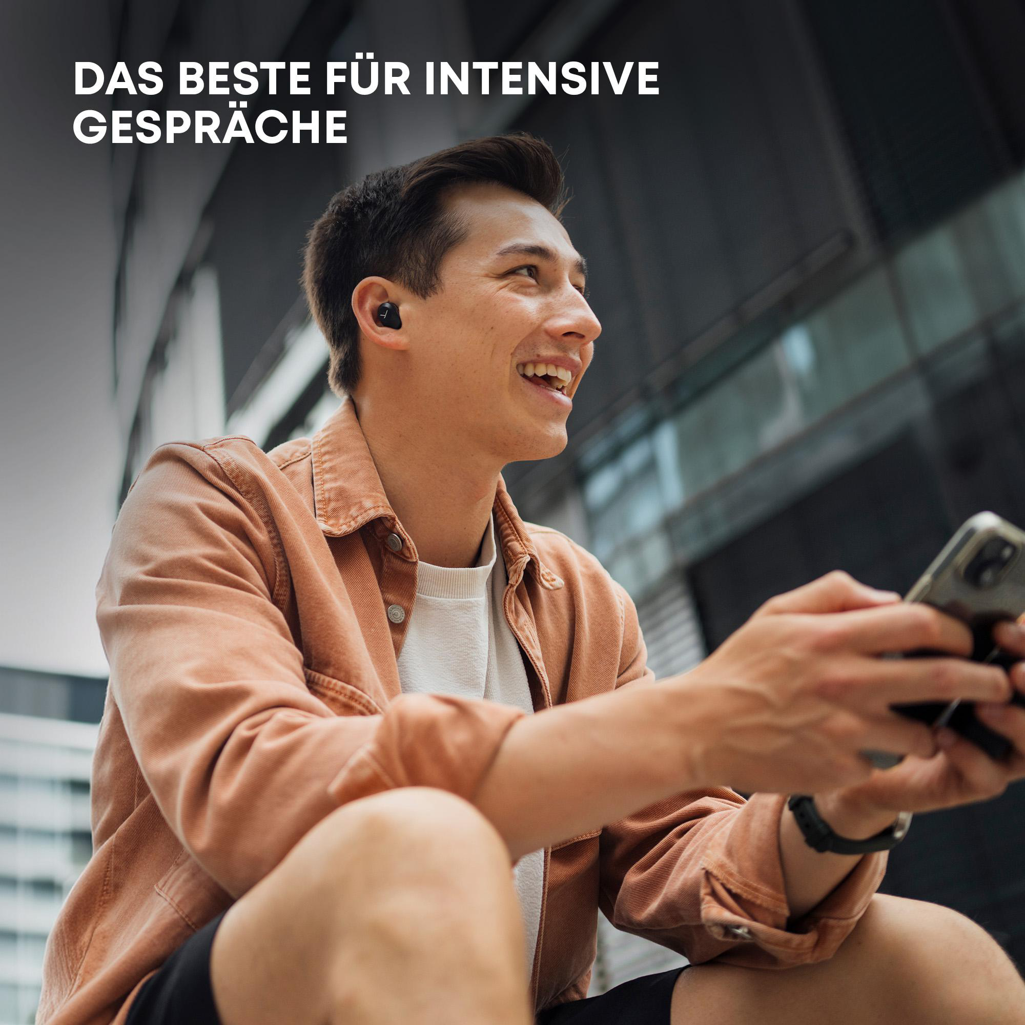 Ein lächelnder Mann mit Ohrhörern hält ein Telefon, mit verschwommenem urbanen Hintergrund.