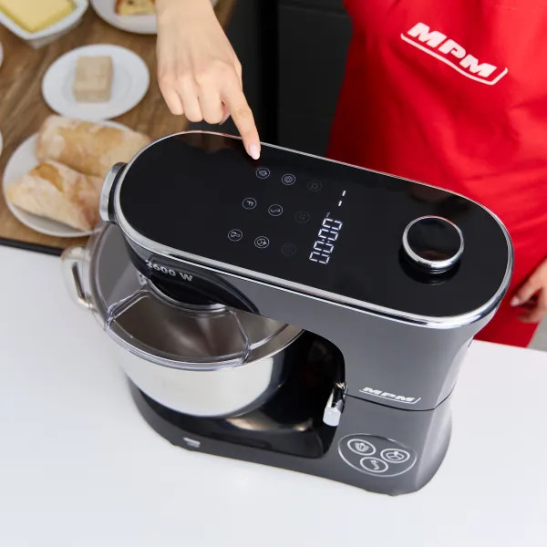 Palec osoby dotyka czarnego urządzenia kuchennego z wyświetlaczem cyfrowym, na białym blacie.