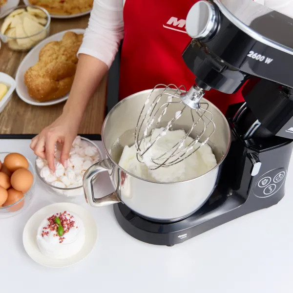 Kobieta robi deser z mikserem stojącym, bezą i jajkami. Różne składniki w tle.