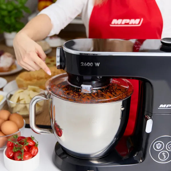 Kobieta wskazuje na czarny mikser stojący. W tle składniki, takie jak jajka i pomidory.