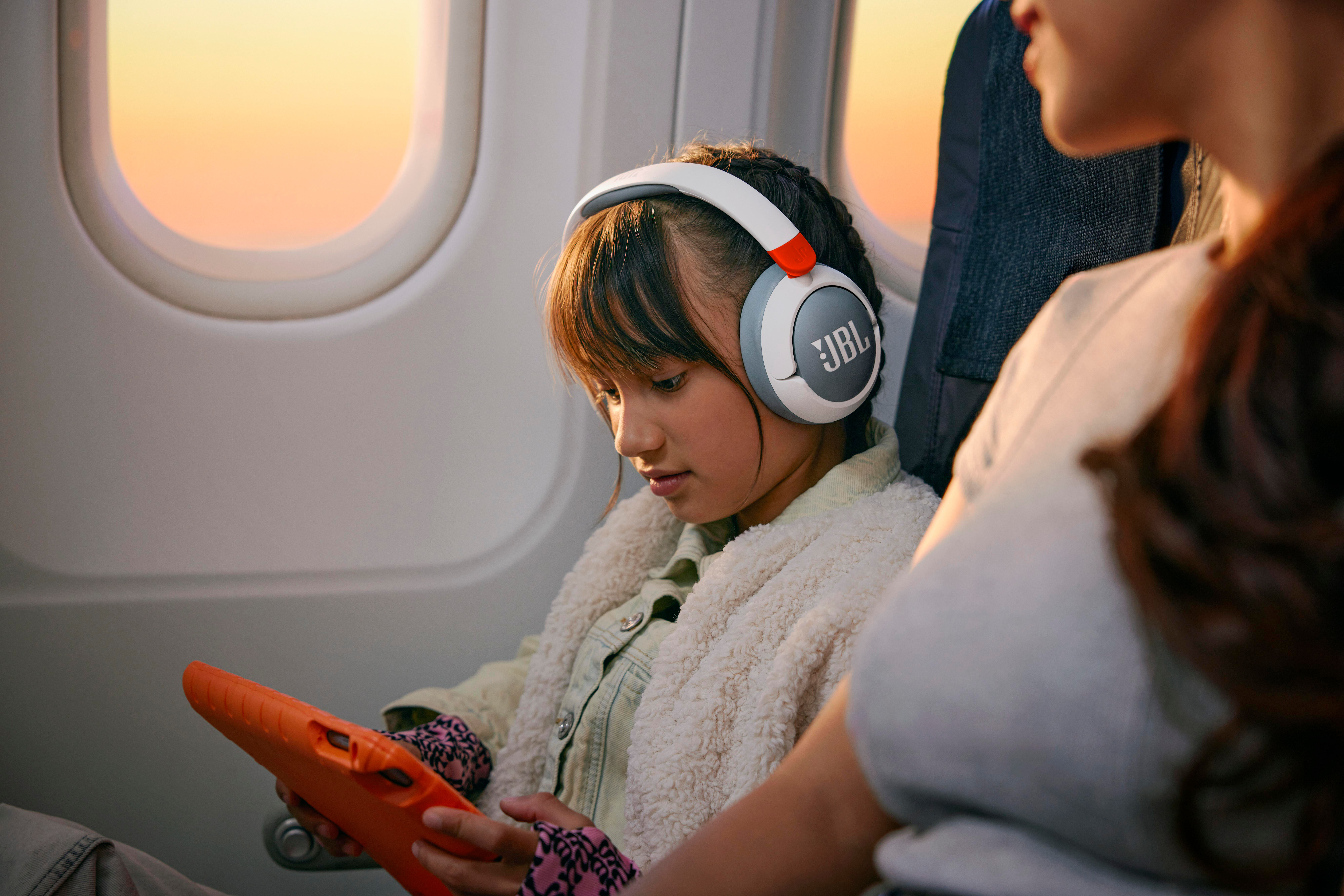 Ein Mädchen mit JBL-Kopfhörern im Flugzeug, das auf ein Tablet schaut.